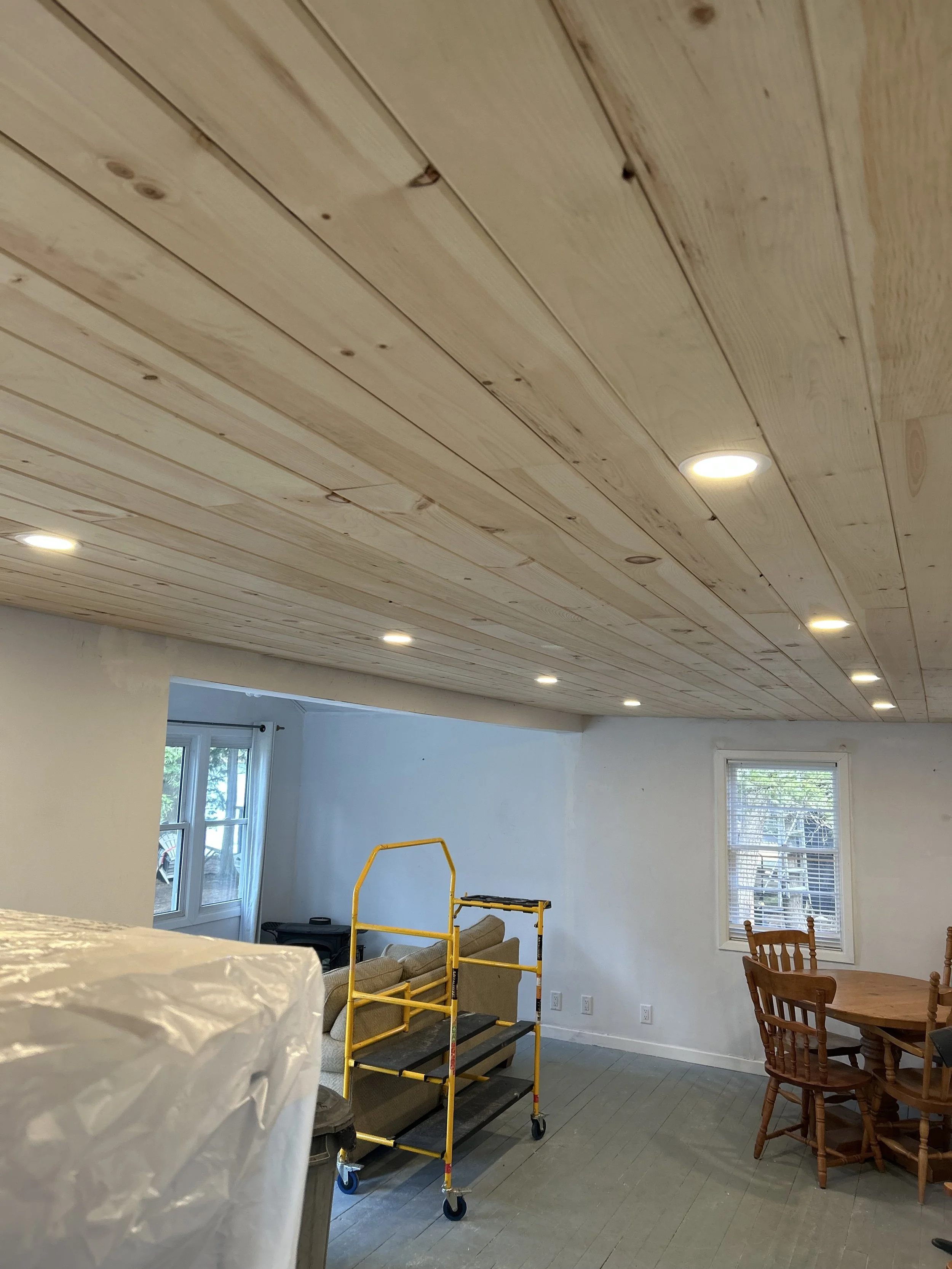 A room with a wooden ceiling, recessed lighting, and a window on the wall. There is a rolling scaffold in front of a beige couch and a round wooden dining table with wooden chairs.