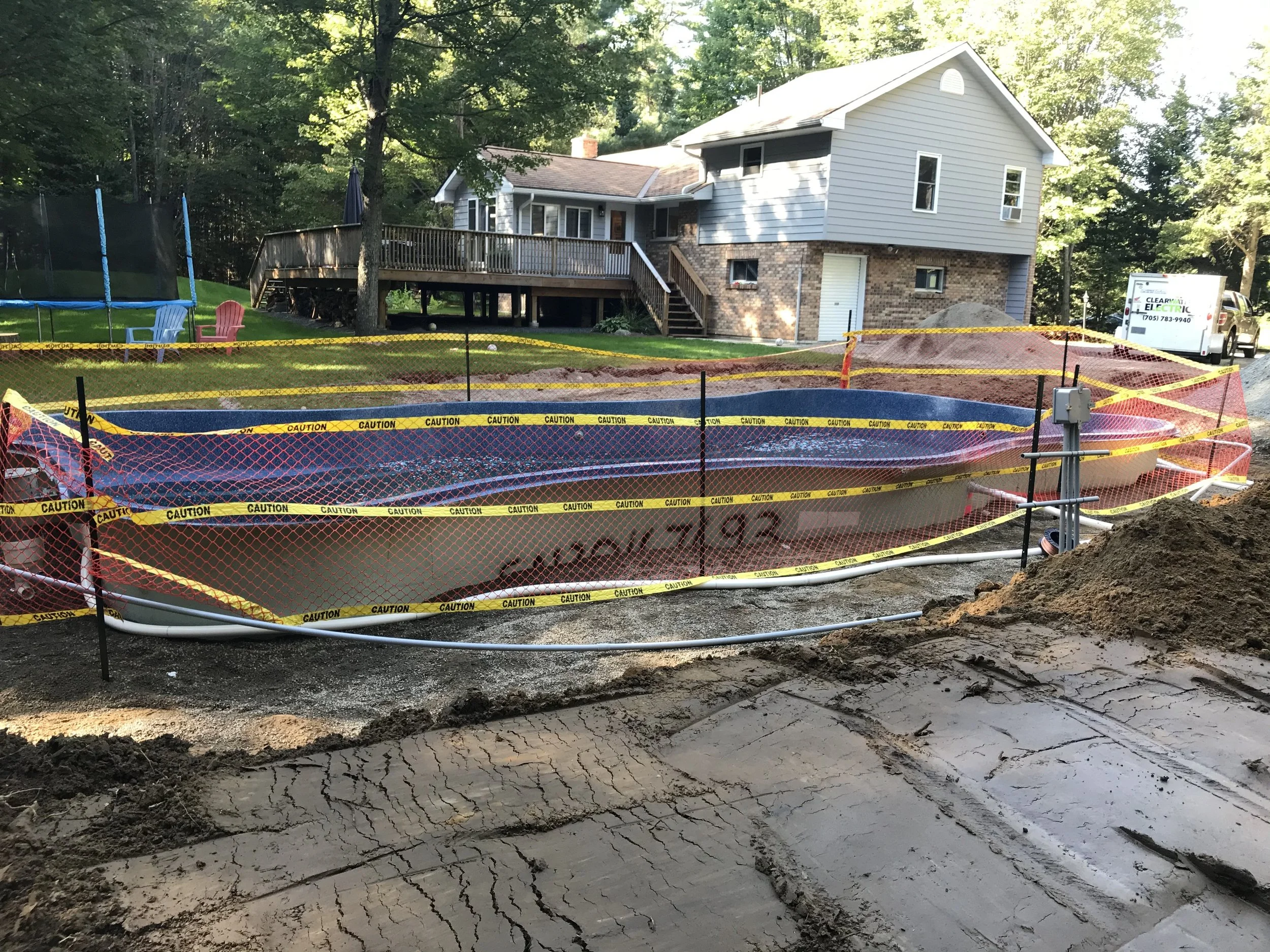 Clearwater Electric installation. A construction site in a backyard with an in-ground pool that is under renovation, surrounded by caution tape and fencing. The backyard has a grassy lawn, trees, and outdoor furniture, with a house in the background.