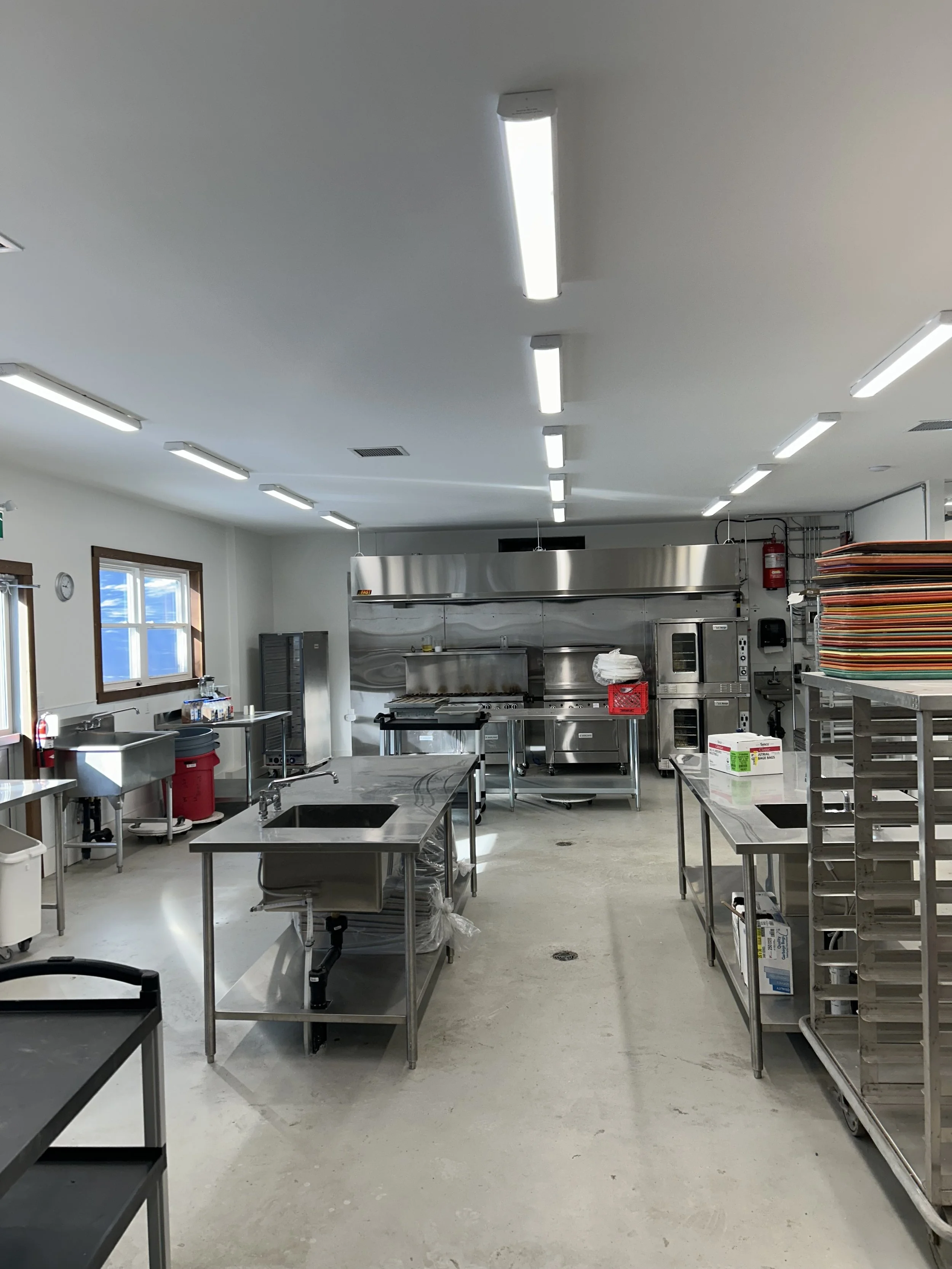 Electrical installation by Clearwater Electric. Industrial kitchen with stainless steel appliances, tables, and racks, illuminated by bright overhead lights.