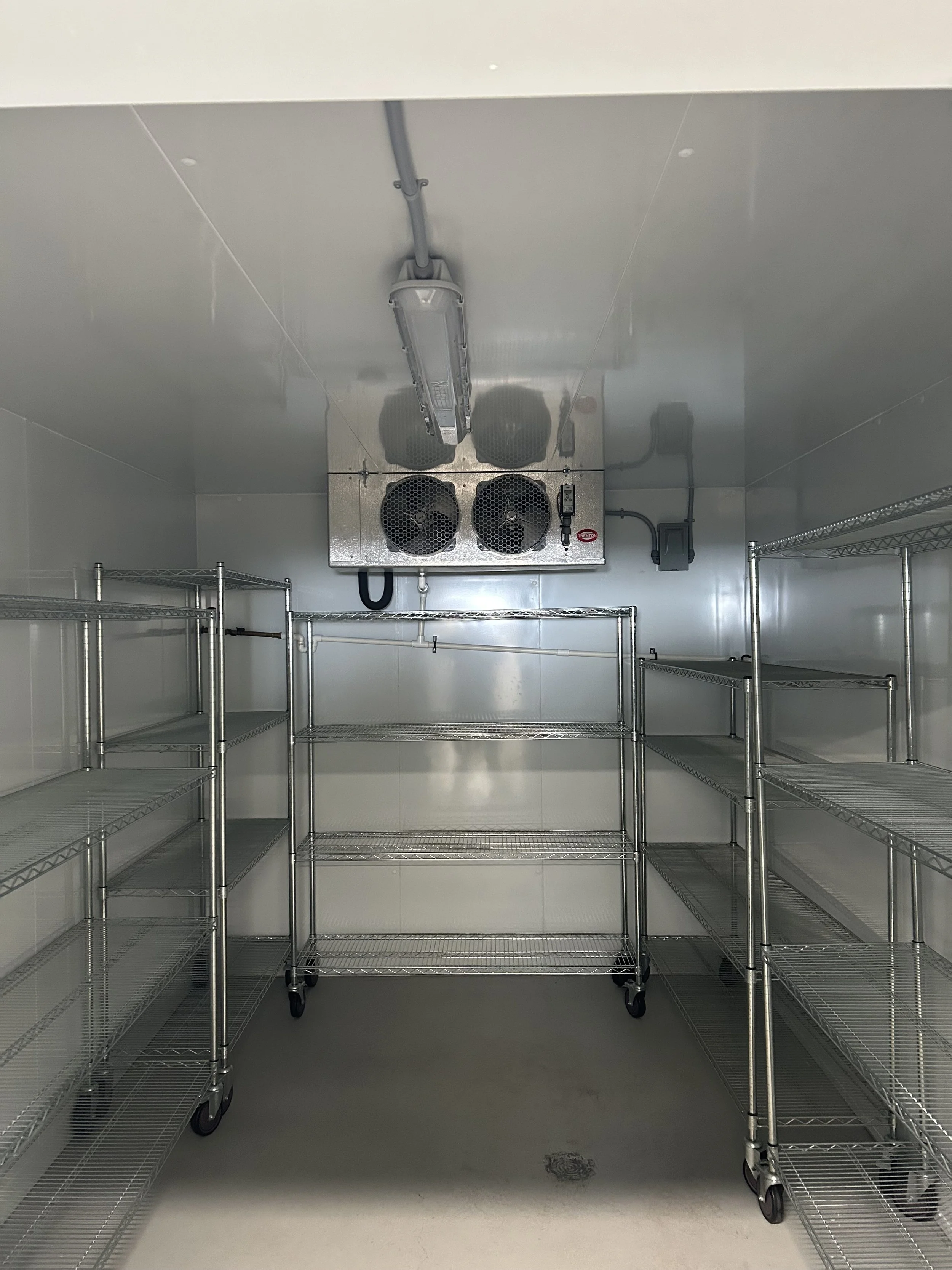 Electrical installation by Clearwater Electric. An empty walk-in refrigeration or storage room with wire shelves and a cooling unit on the ceiling.