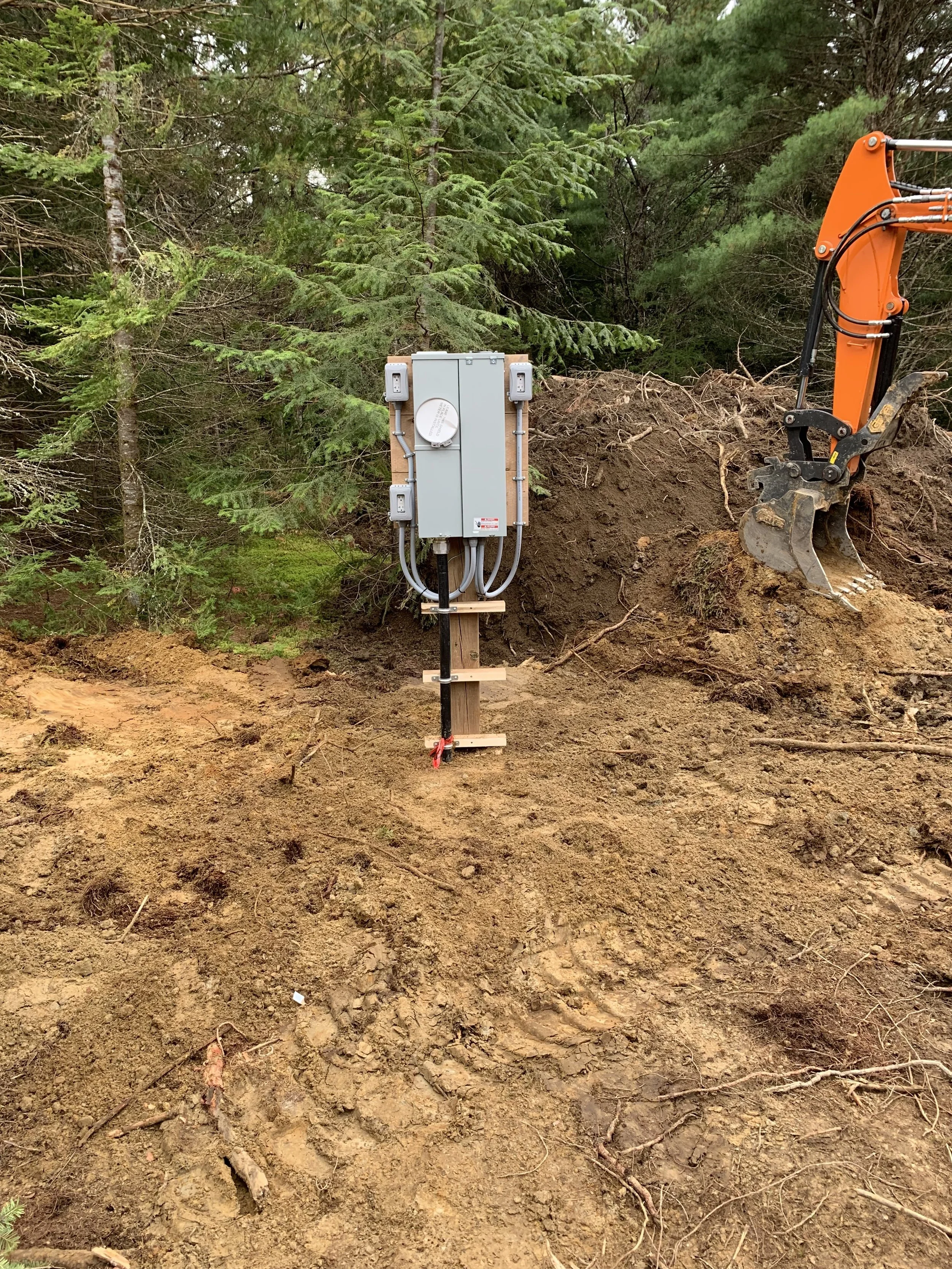 Electrical job by Clearwater Electrical. Electrical transformer box mounted on a wooden stand at a construction site surrounded by dirt and trees, with part of an orange excavator visible on the right.