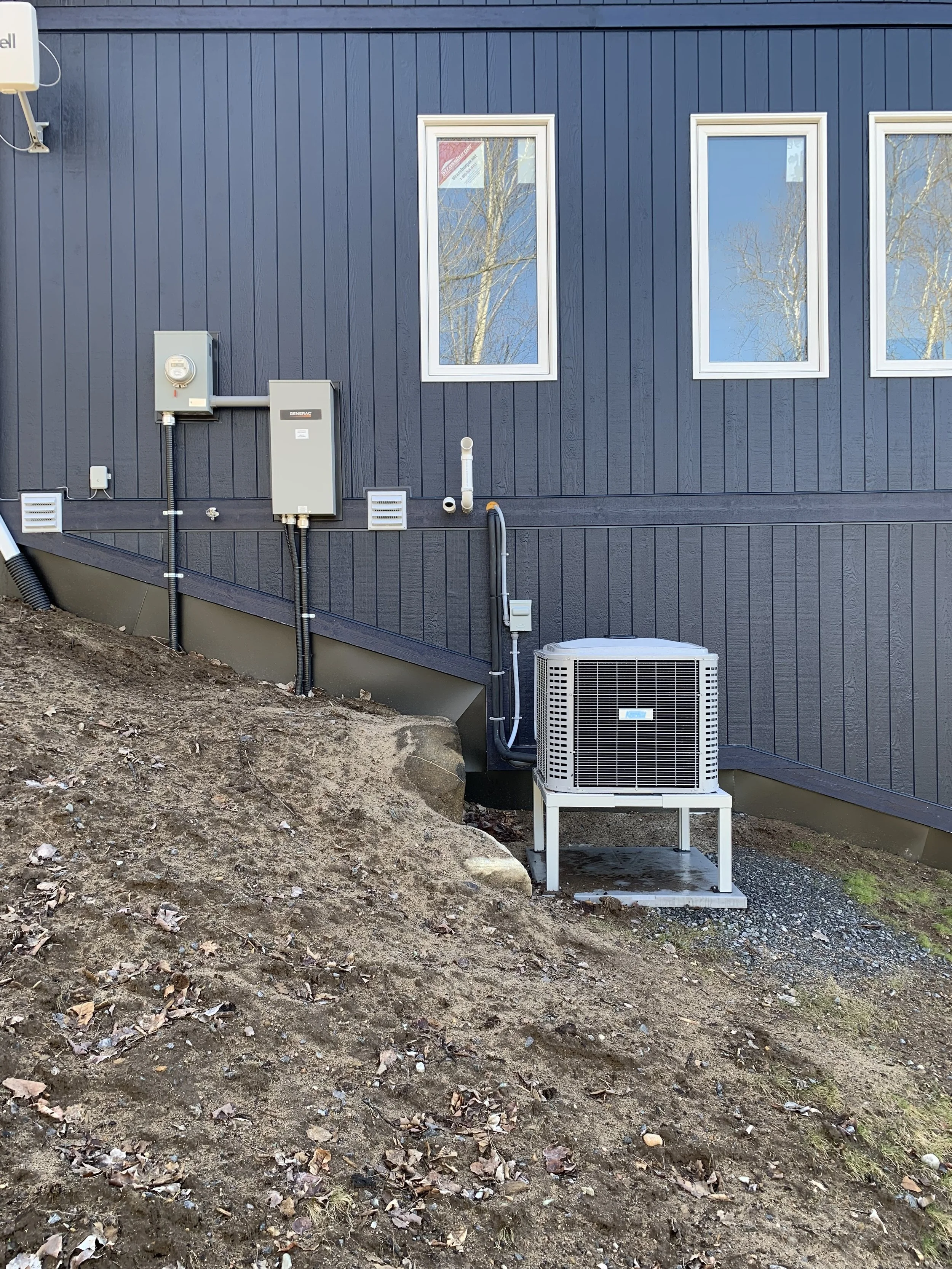 Electrical installation by Clearwater Electric. Exterior view of a blue house with three windows and an HVAC unit outside, placed on a metal stand, surrounded by dirt and fallen leaves.