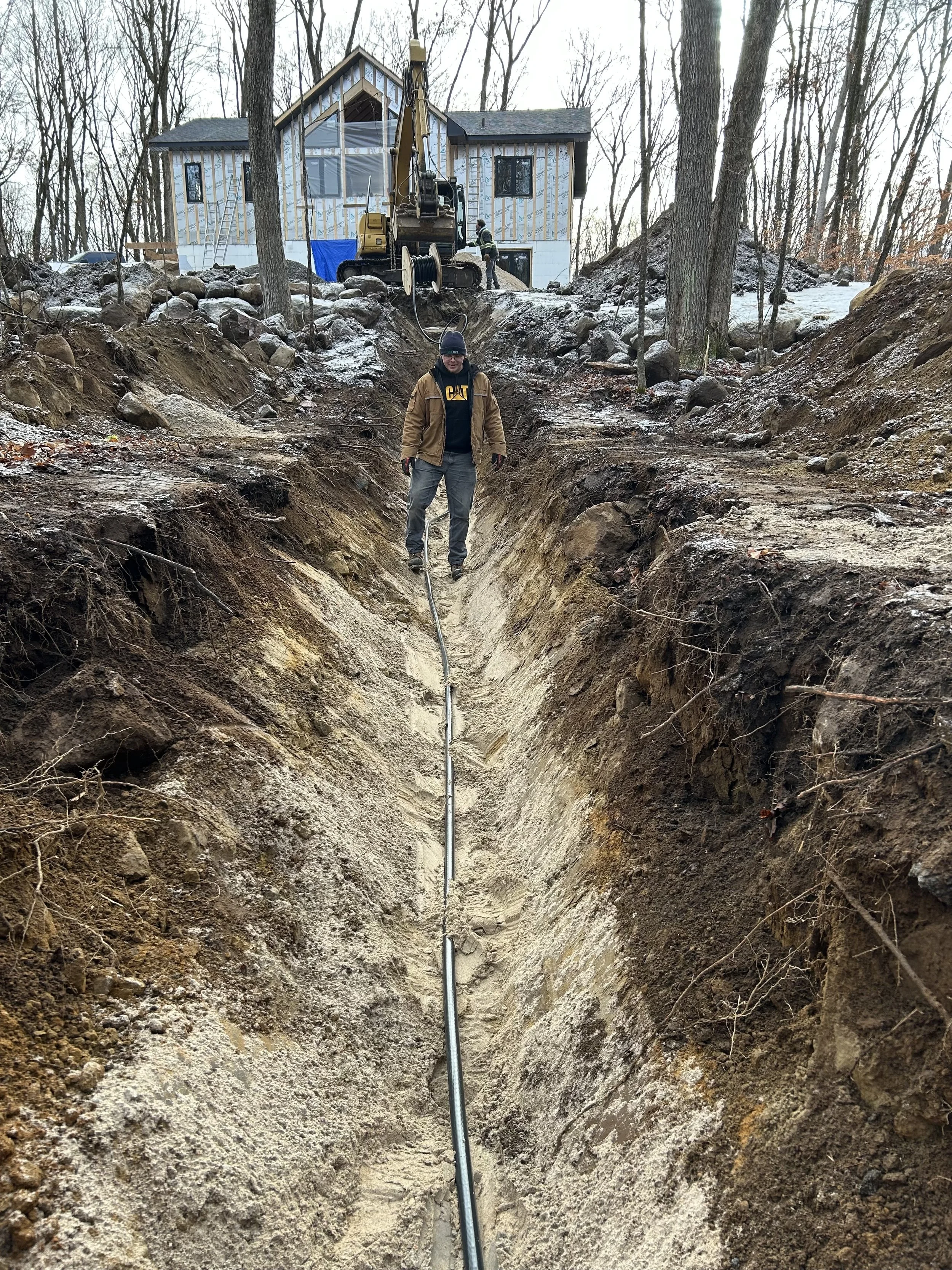 A person standing in a deep trench during a construction project, with a house and construction equipment visible in the background.