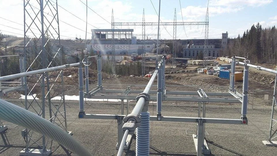 View of a construction site with power lines and electrical infrastructure in the foreground, buildings, and forests in the background.