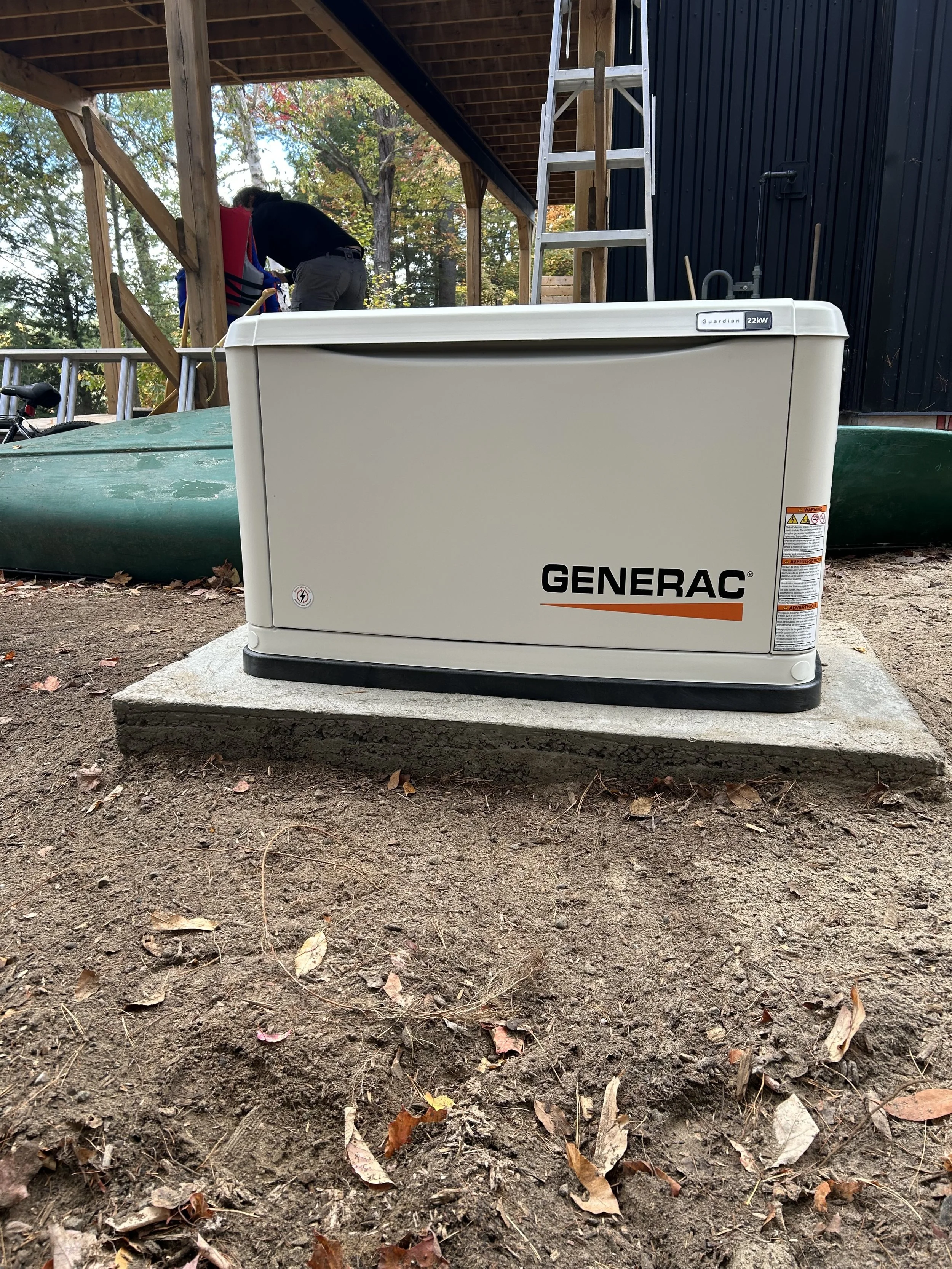A Generac standby generator installed outdoors on a concrete pad on dirt ground, with a wooded area and a wooden structure in the background.