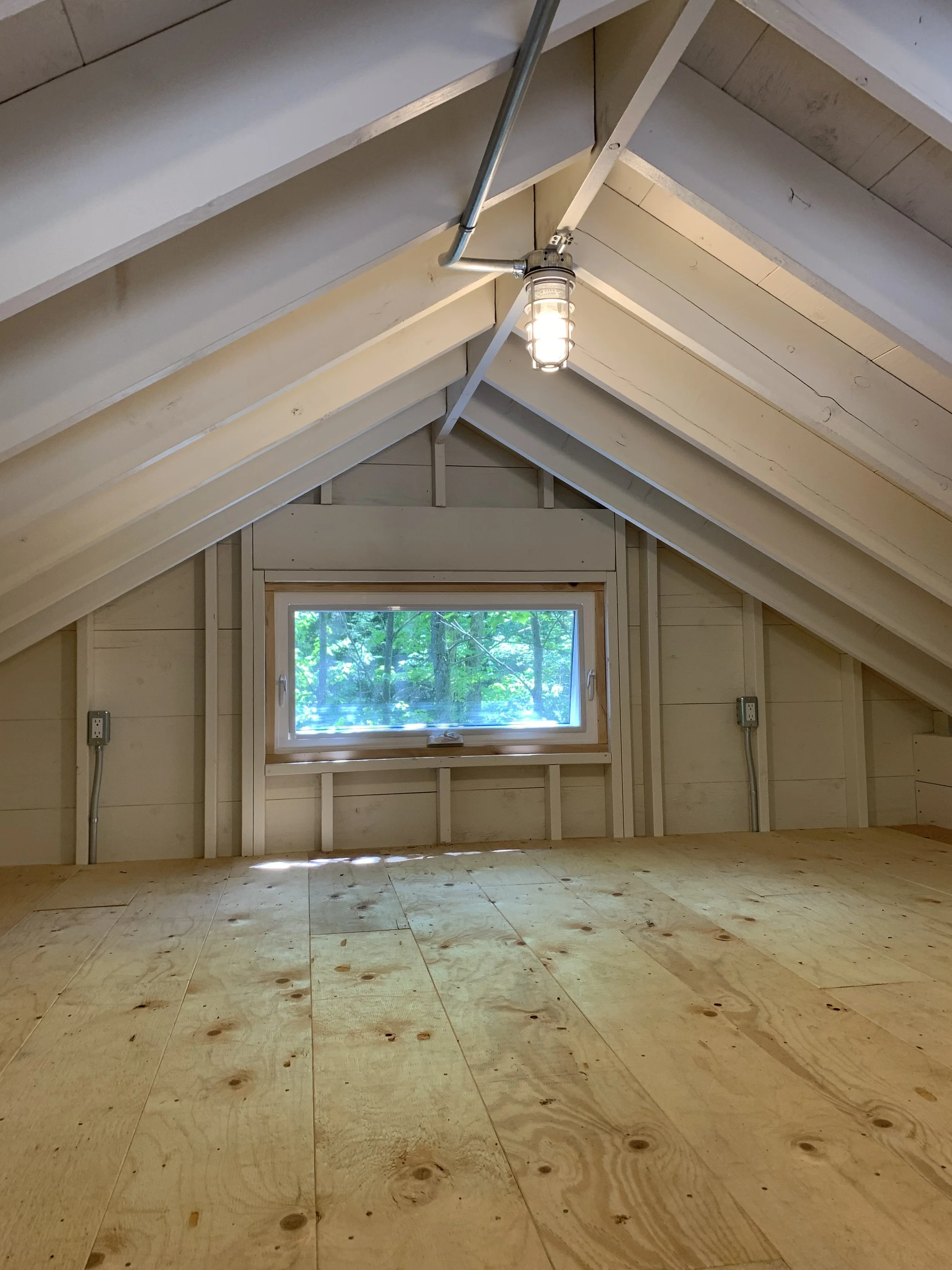 Electrical installation by Clearwater Electric. Empty unfinished attic with plywood floor, small window, and ceiling light fixture.
