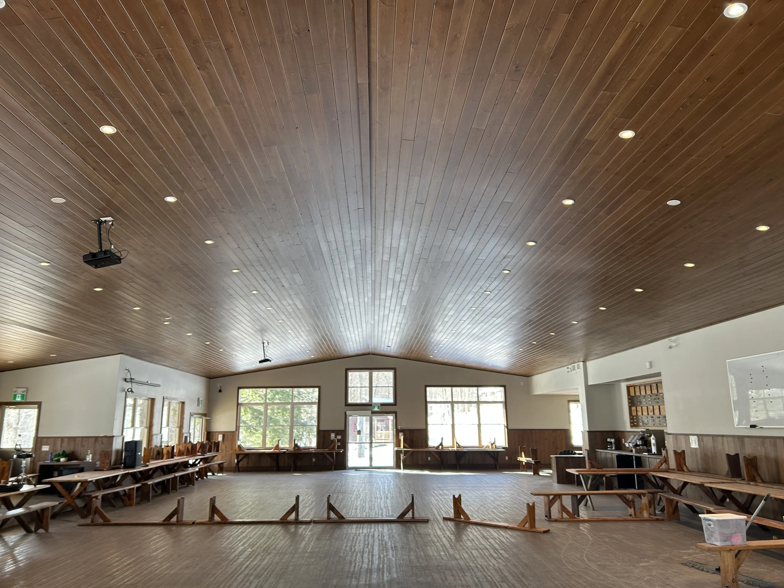 Electrical installation by Clearwater Electric. Large indoor room with a wooden ceiling and multiple windows, tables along the walls, and a light-colored carpeted floor, likely a community or event space.
