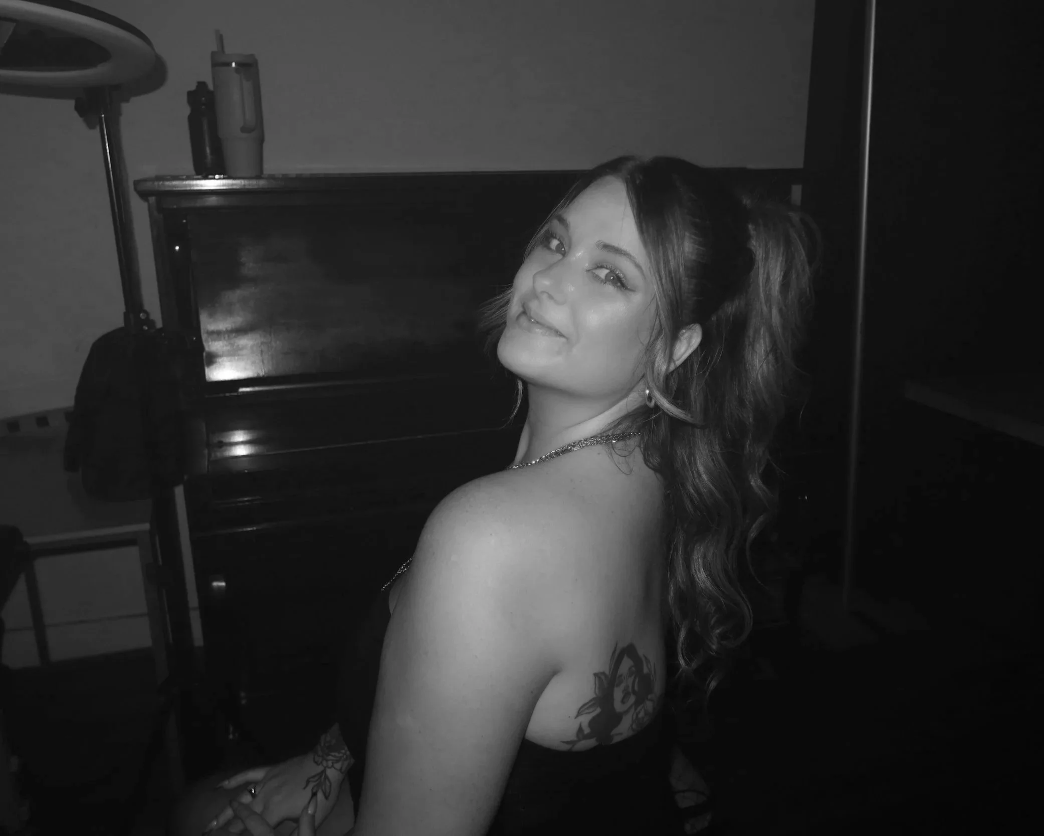 Une femme souriante avec des cheveux bouclés portant une robe noire, assise devant un piano et affichant un tatouage d'une araignée sur le bras gauche.