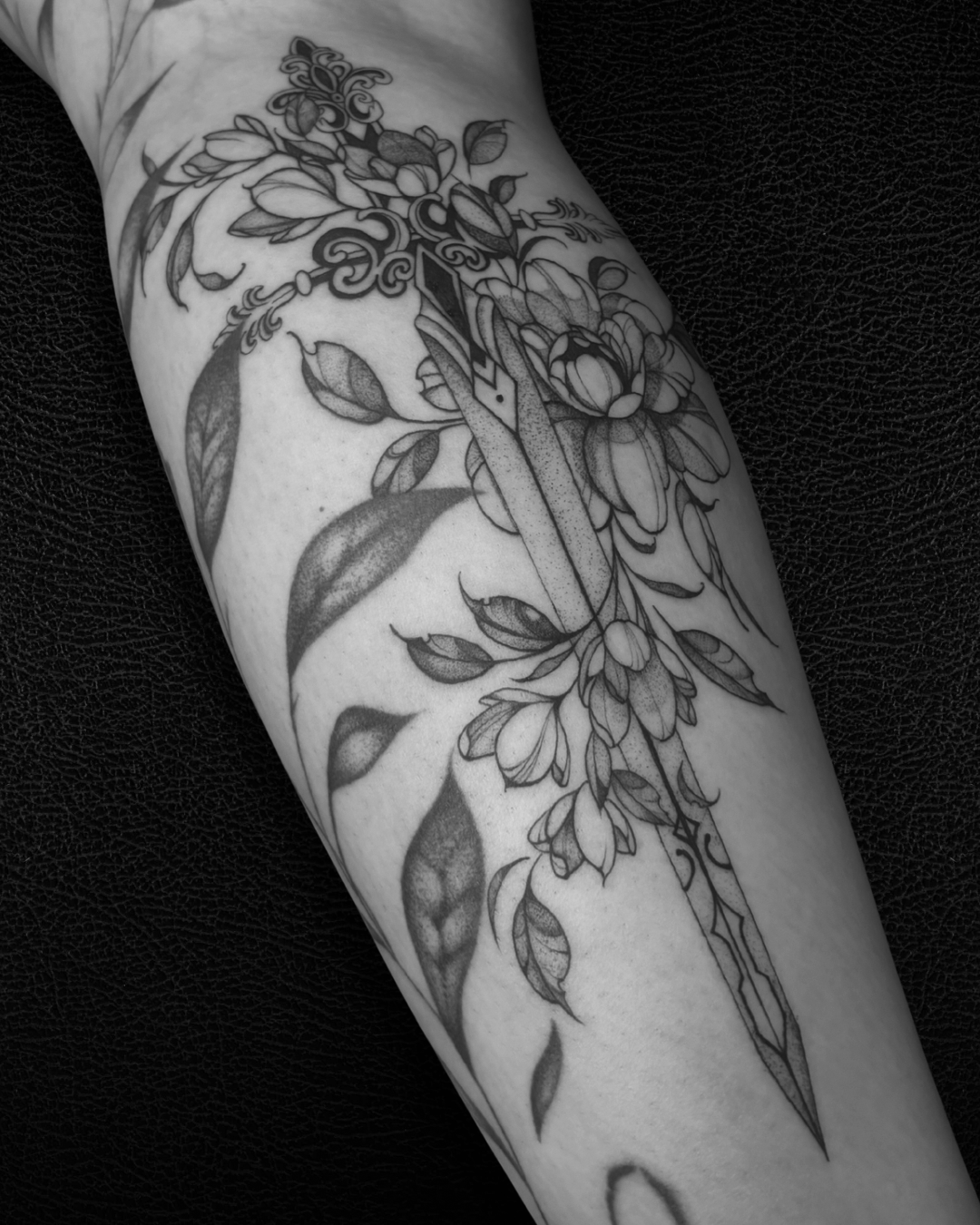 Tatouage noir et blanc d'un croisement d'éléments floraux et d'une épée, avec des feuilles et des fleurs