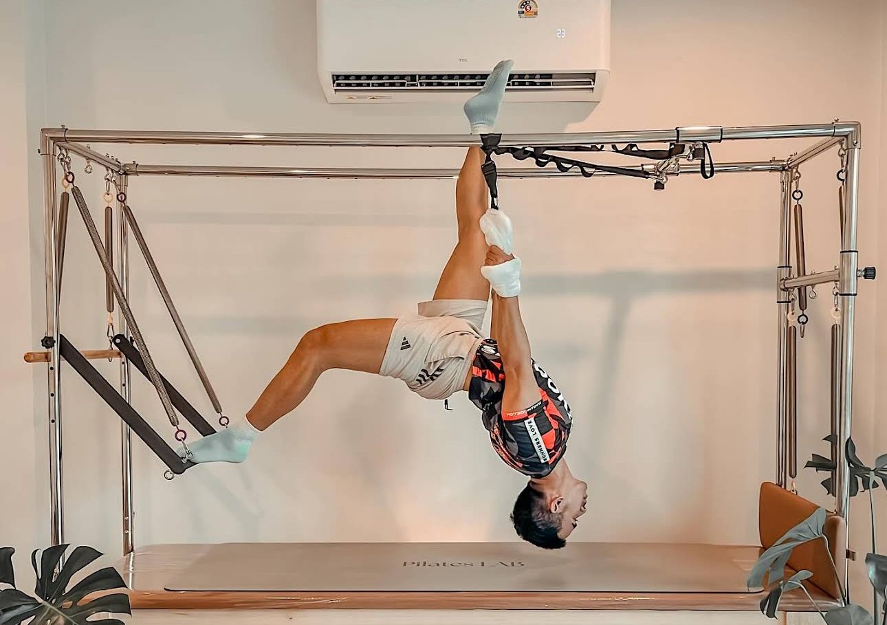 A man performing an aerial pilates pose using suspension straps in a studio with a metal frame, an air conditioning unit above, a yoga mat on the floor, and plants on the sides.