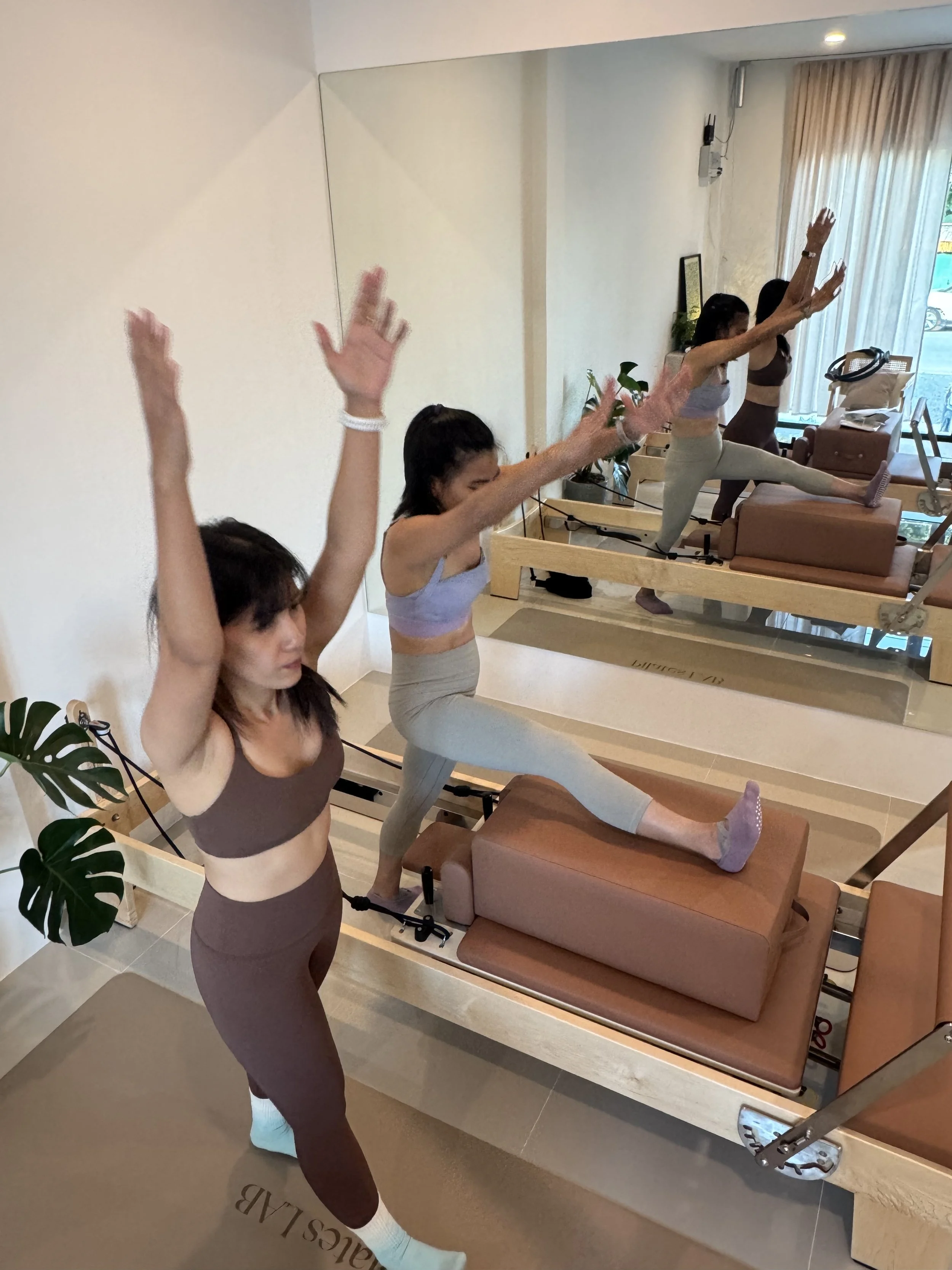 Two women practicing pilates in a fitness studio, performing a balancing pose on yoga raised mats in front of a large mirror.