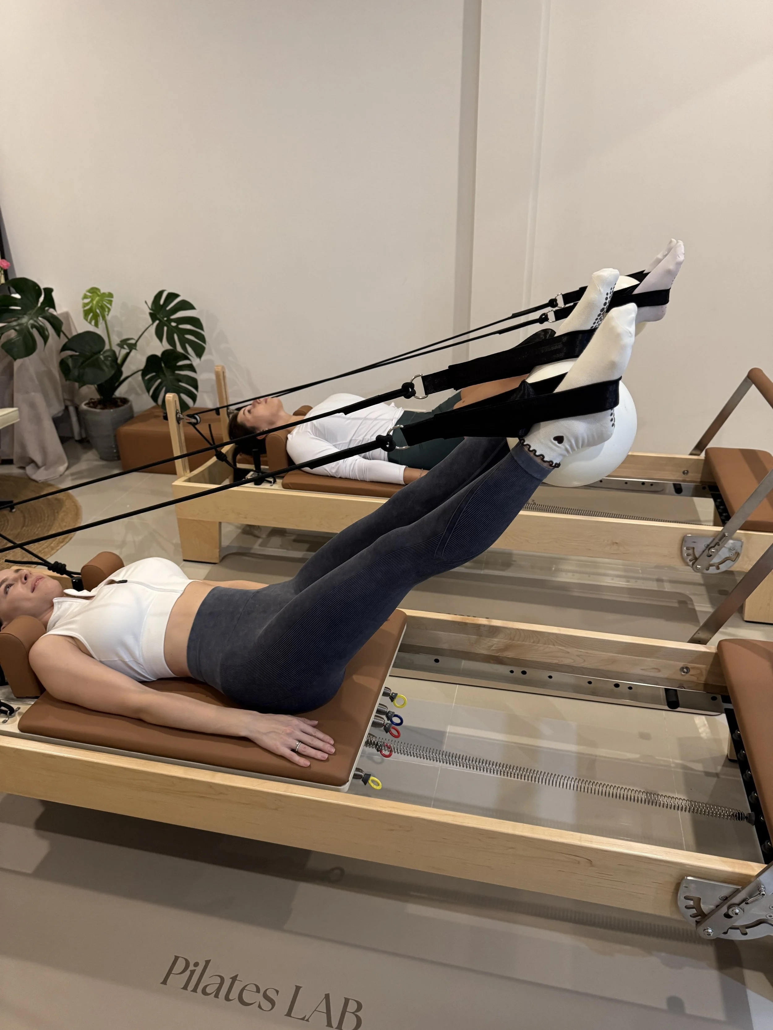 Two women lying on a Pilates reformer machine performing ab exercises.