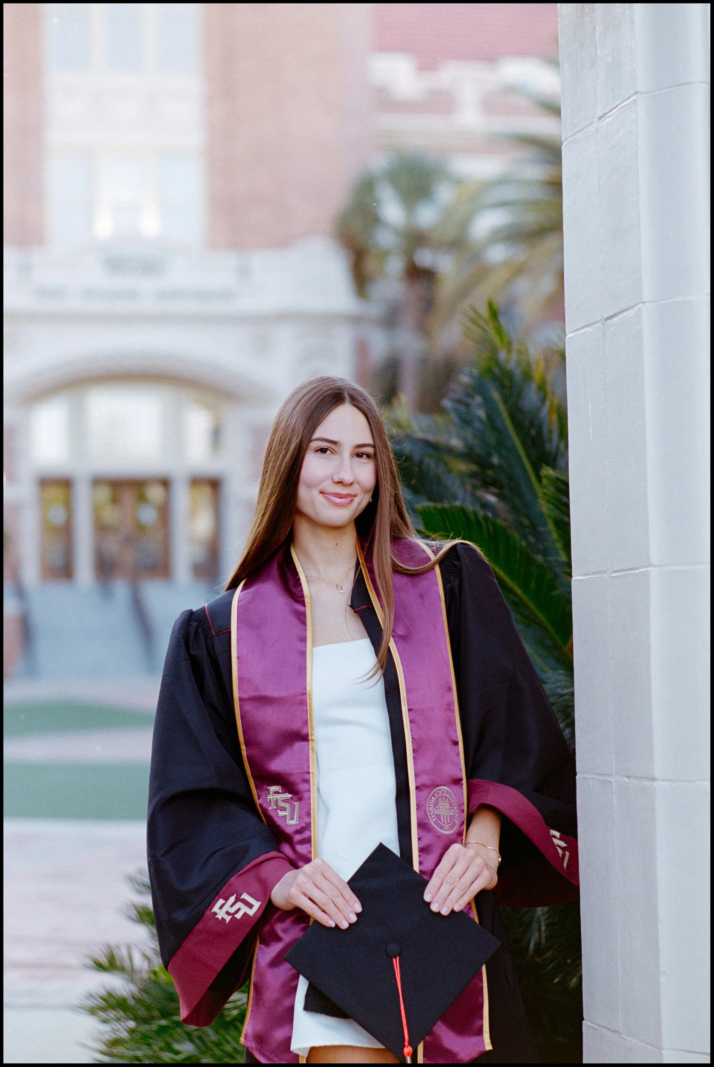 A young woman in graduation attire, holding a cap, standing outdoors with a campus background, smiling.