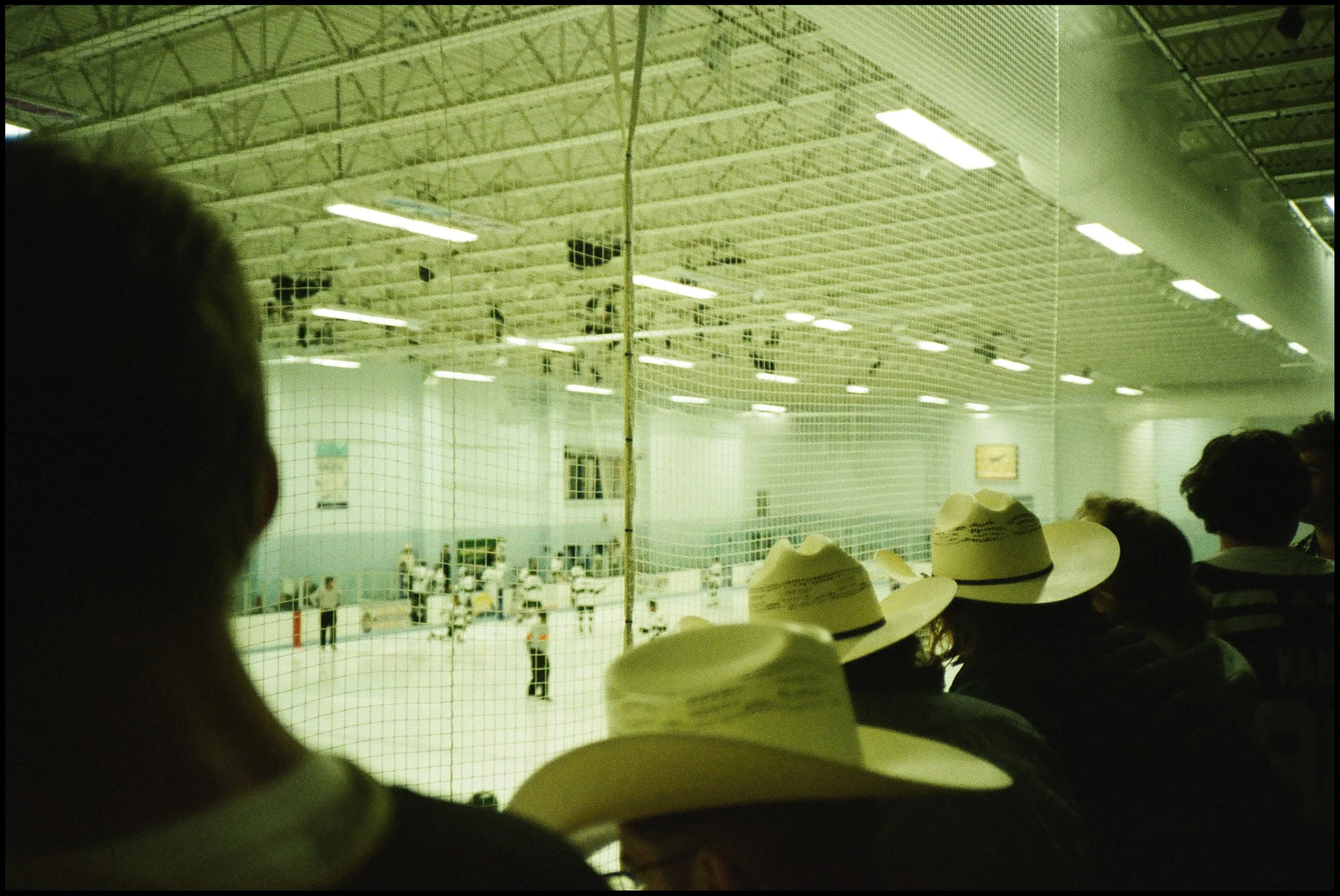 Ice hockey game viewed through a protective glass in an indoor rink with spectators wearing cowboy hats in the foreground.