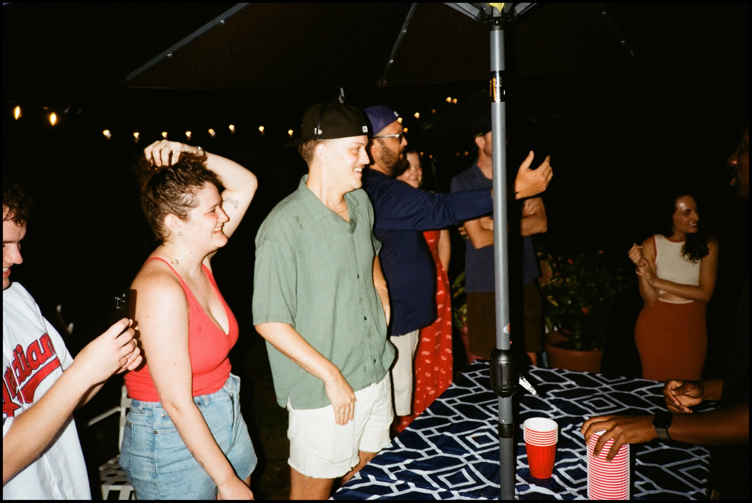 People gathered around a table at night, celebrating under a large umbrella with string lights. One woman in a red top and denim shorts is smiling, while a man in a green shirt and cap is also smiling. Others are holding red cups, engaging in conversation, with a woman wearing a red dress in the background.