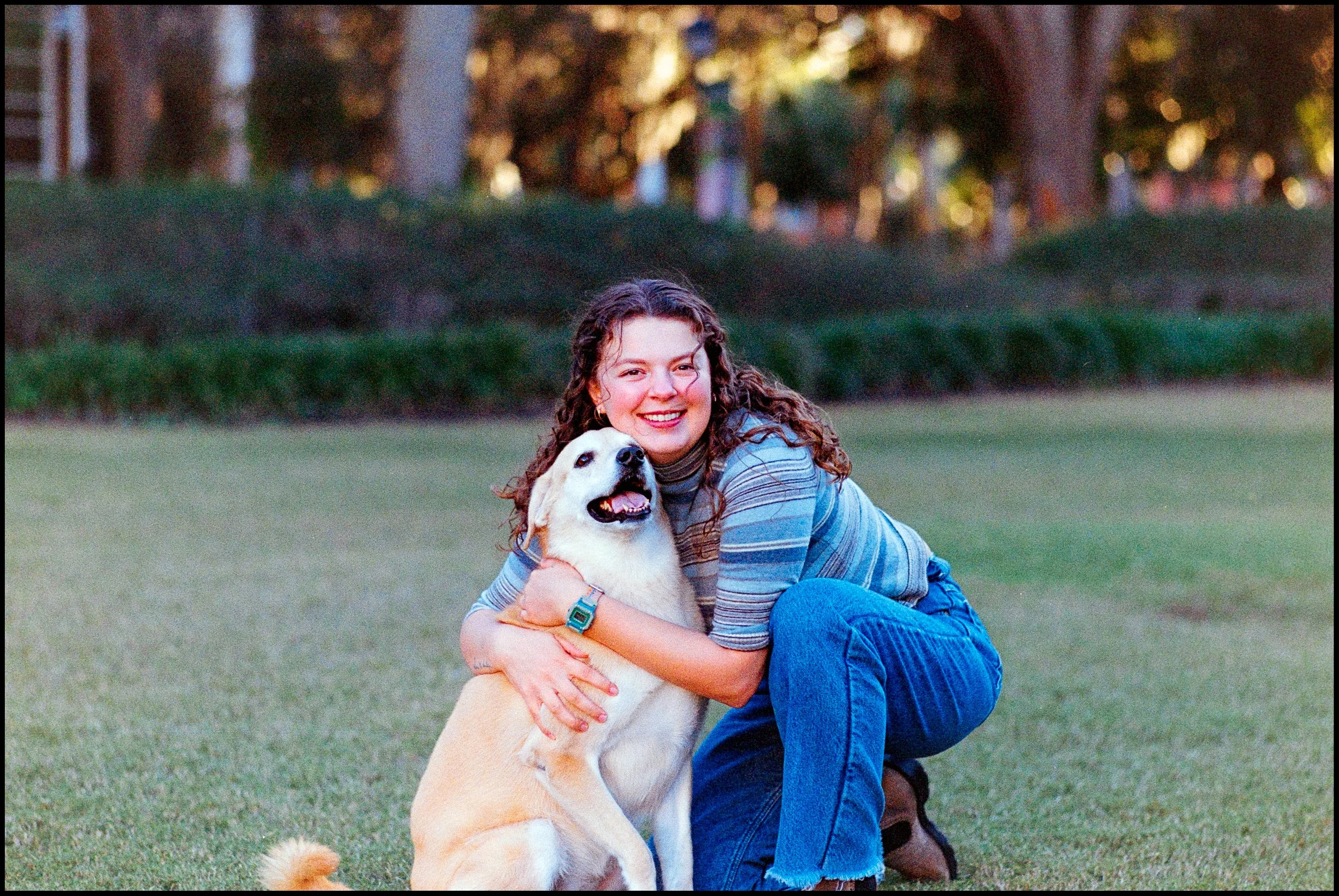 A young woman with curly hair, wearing a striped long-sleeve shirt and blue jeans, kneeling on the grass and hugging a big, light-colored dog with a happy expression, in a park-like setting with trees in the background during daytime.