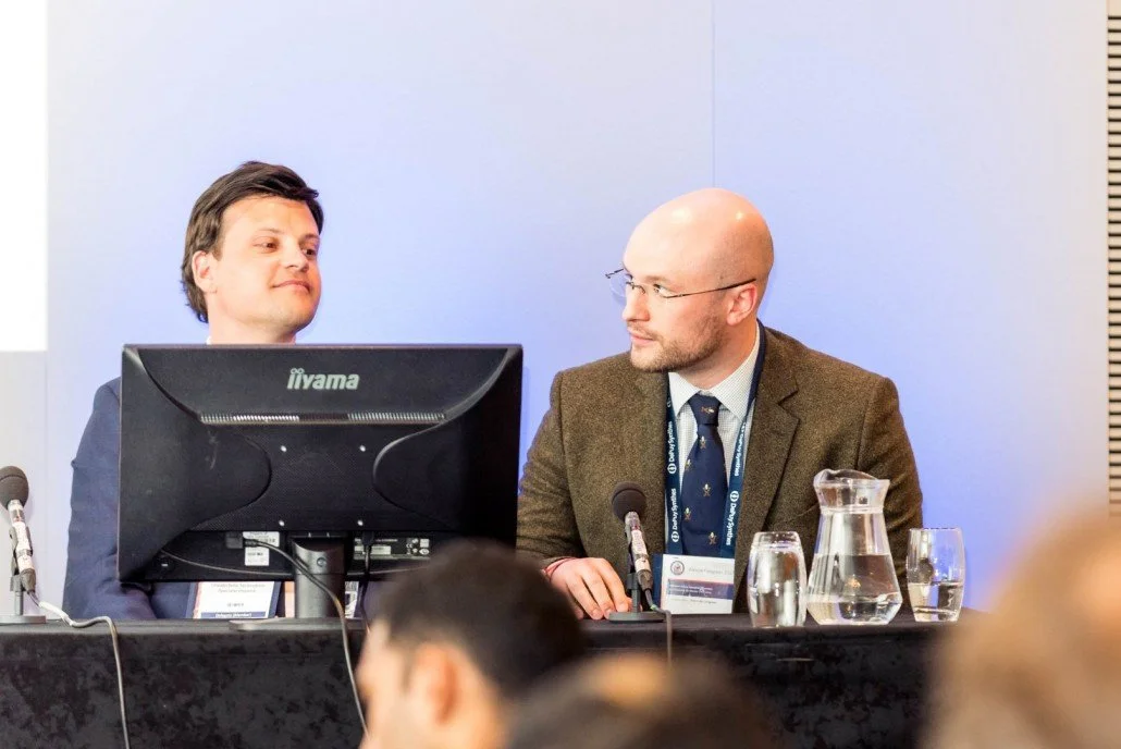 Two men sitting at a conference table, one facing a monitor and the other looking at him, with microphones and glasses of water in front of them.