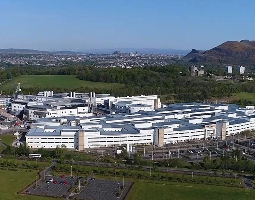A large hospital complex with white buildings is shown in the foreground, surrounded by parking lots and green landscaping. In the background, there are open fields, forests, and a city skyline with hills and mountains in the distance under a clear blue sky.
