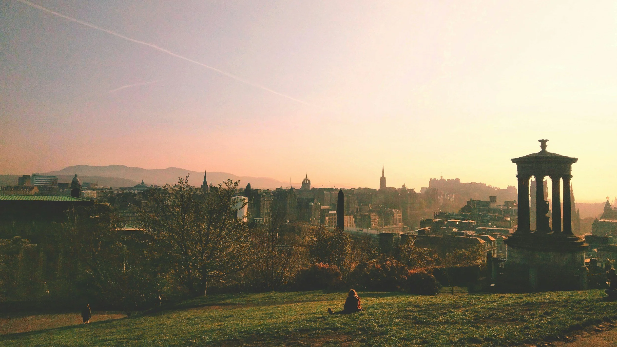 A cityscape view during sunset with a park in the foreground, a person sitting on the grass, trees, historic buildings, and a monument with columns.