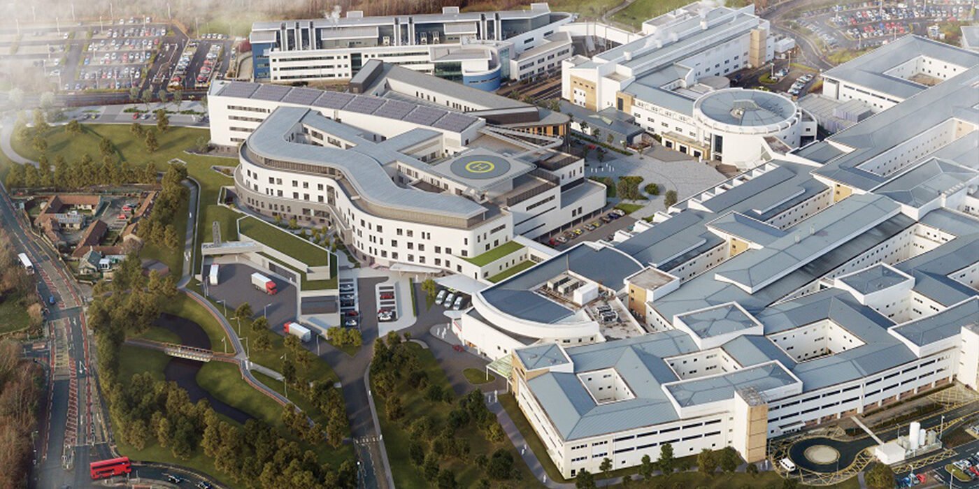 Aerial view of a large modern hospital complex with multiple interconnected white buildings, parking lots, green spaces, and a helipad on the roof.