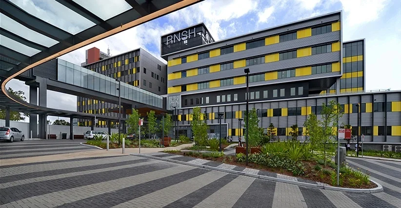 Modern hospital building with yellow and gray exterior, parking lot, trees, and clear sky.
