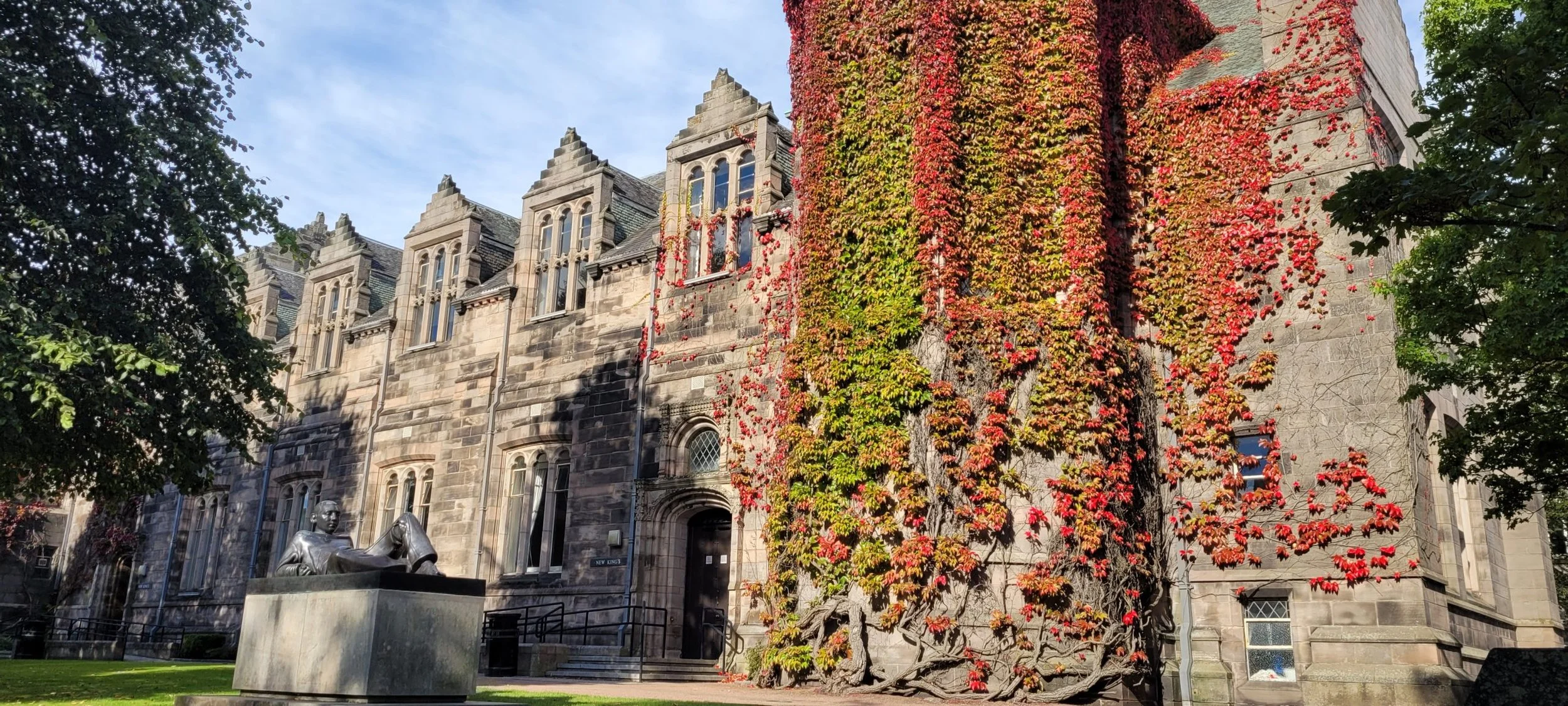 A historic stone building with gothic windows, partially covered in colorful autumn leaves, with a statue of a reclining person in front, and trees casting shadows on the facade.