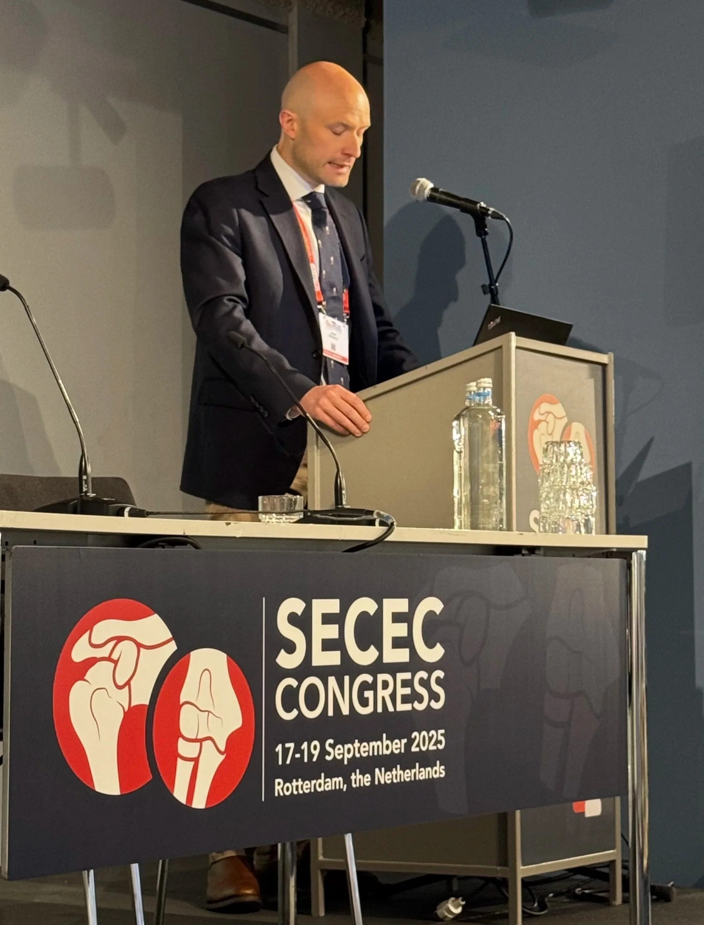 A man in a dark suit and tie standing at a podium delivering a speech at the SECEC Congress held from September 17 to 19, 2025, in Rotterdam, the Netherlands. The podium has a sign with the event details and a logo featuring two hands.