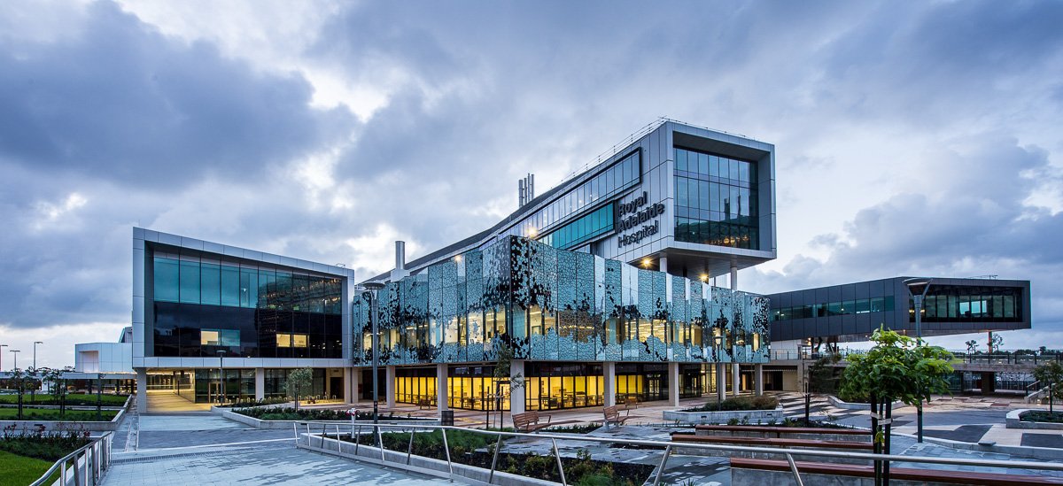 Modern hospital building with glass windows and geometric design, labeled as Royal Adelaide Hospital, with outdoor walkways and landscaping, under a cloudy sky.