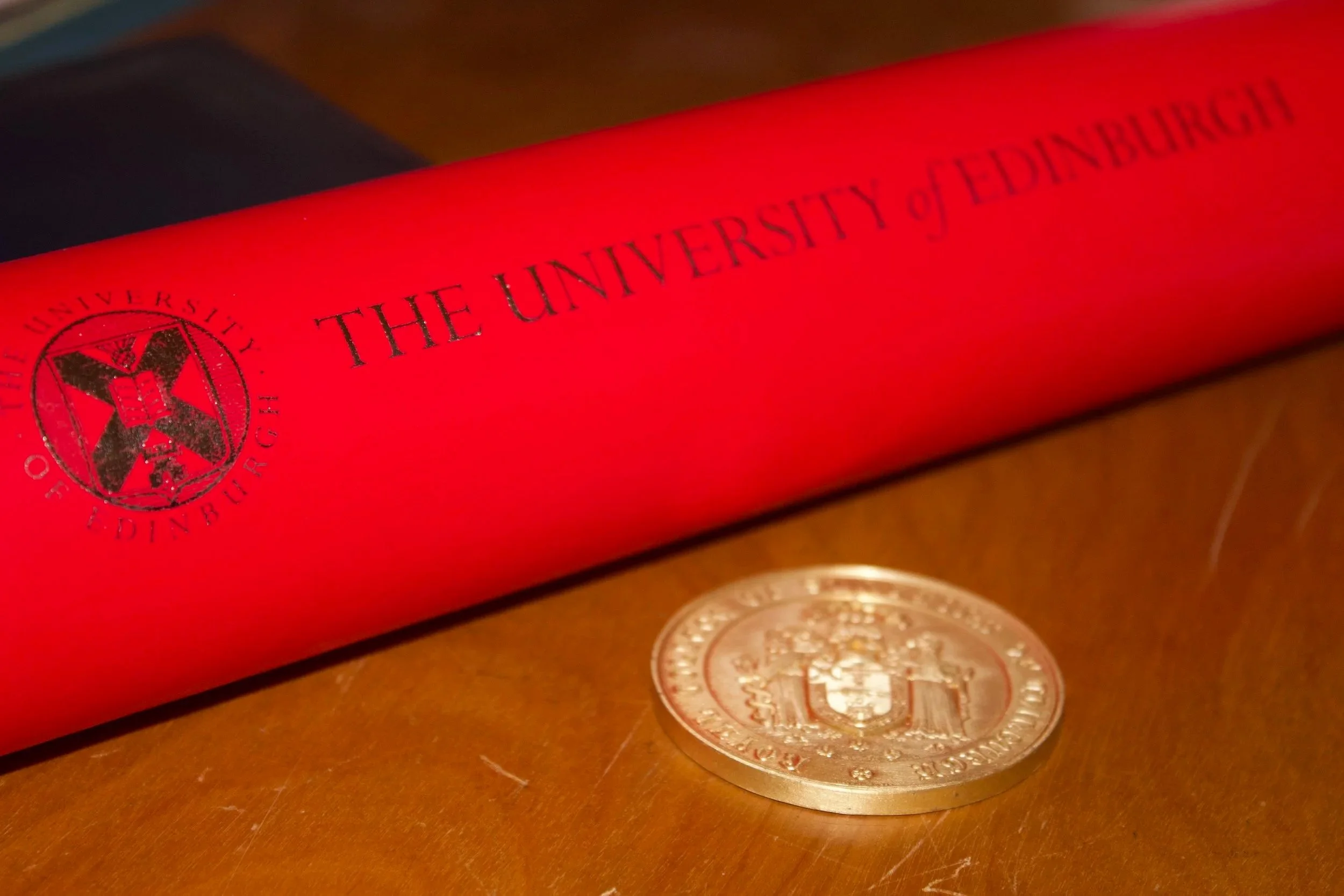 Red diploma cover from the University of Edinburgh with a gold coin in front of it.