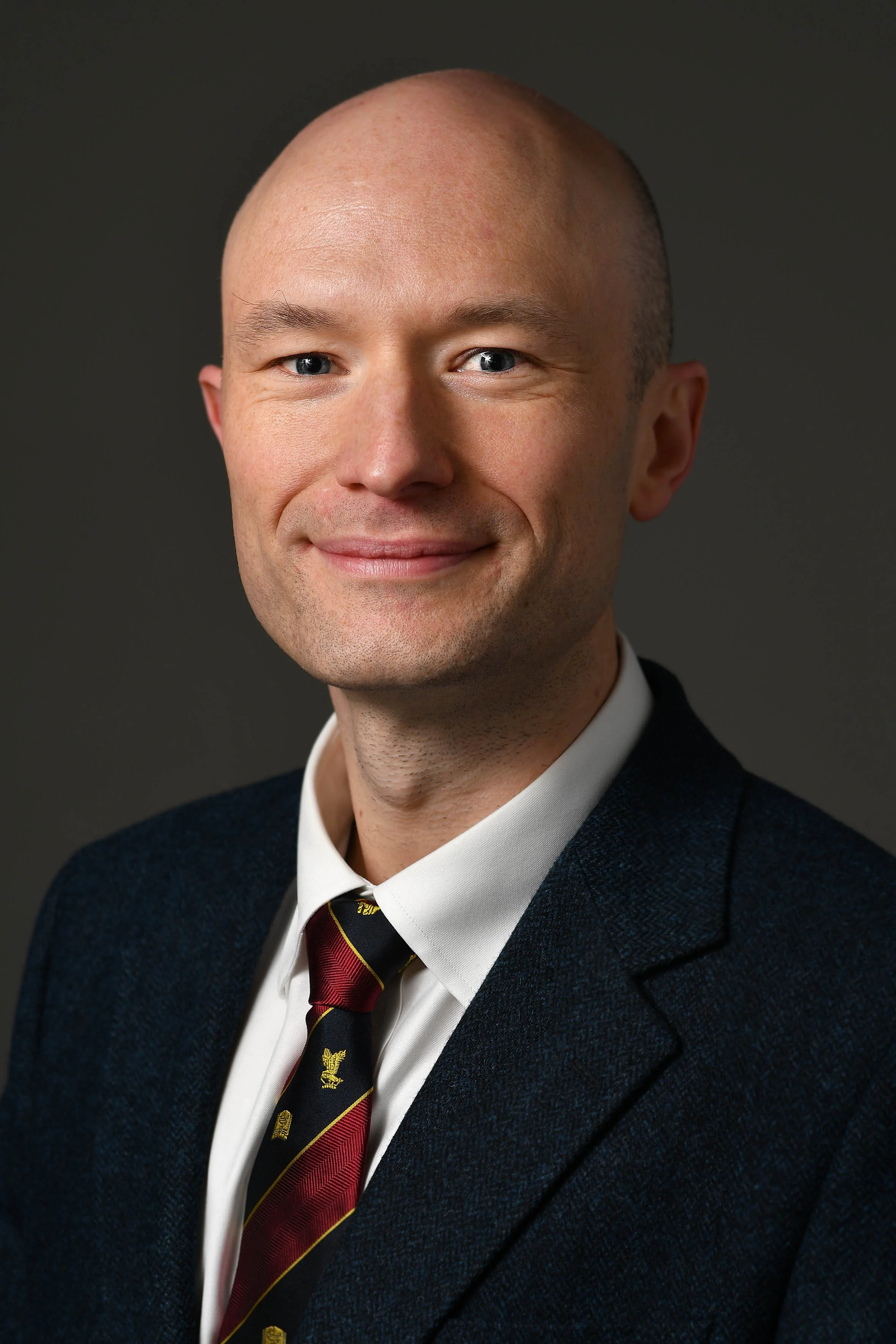 A professional headshot of a bald man in a suit and tie, smiling at the camera.