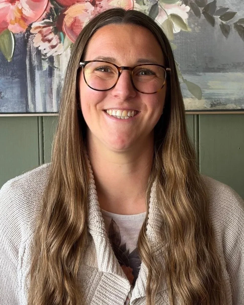 A woman with long brown hair and glasses, smiling in front of a floral painting on a green wall.