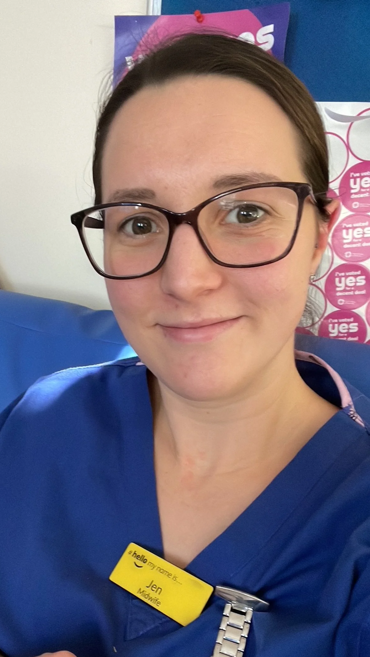Close-up of a woman with glasses, wearing a blue uniform with a name tag that says Jen, sitting on a blue chair in what appears to be a healthcare or retail setting.