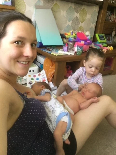 A woman sitting on the floor with two young children, possibly in an indoor playroom or living room. One child is sitting on her lap, and the other is sitting nearby. The background includes toys, books, and children's items on a wooden table.