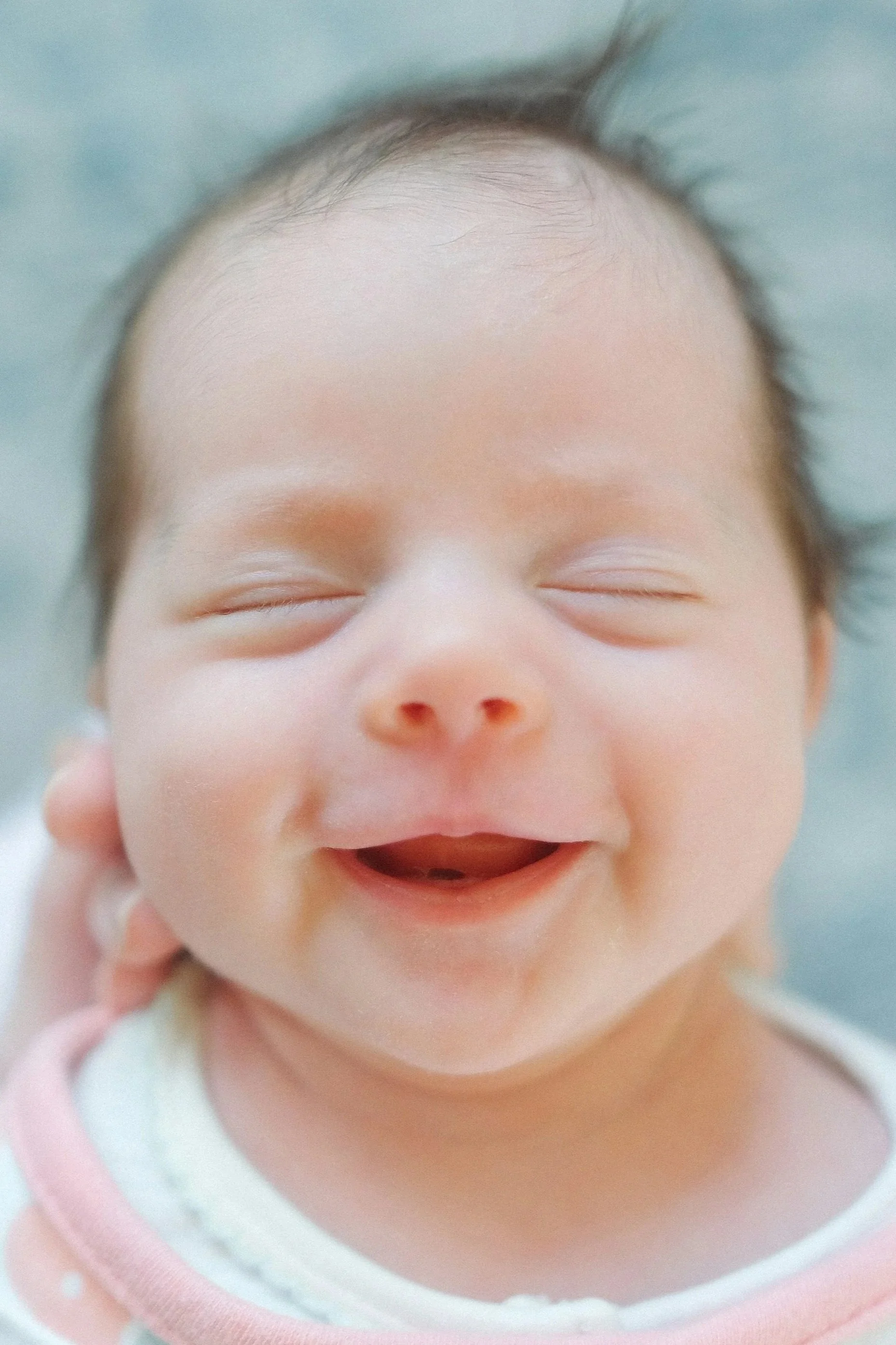 Close-up of a laughing baby's face with eyes closed and mouth open, showing baby fat cheeks and fine hair on the forehead.