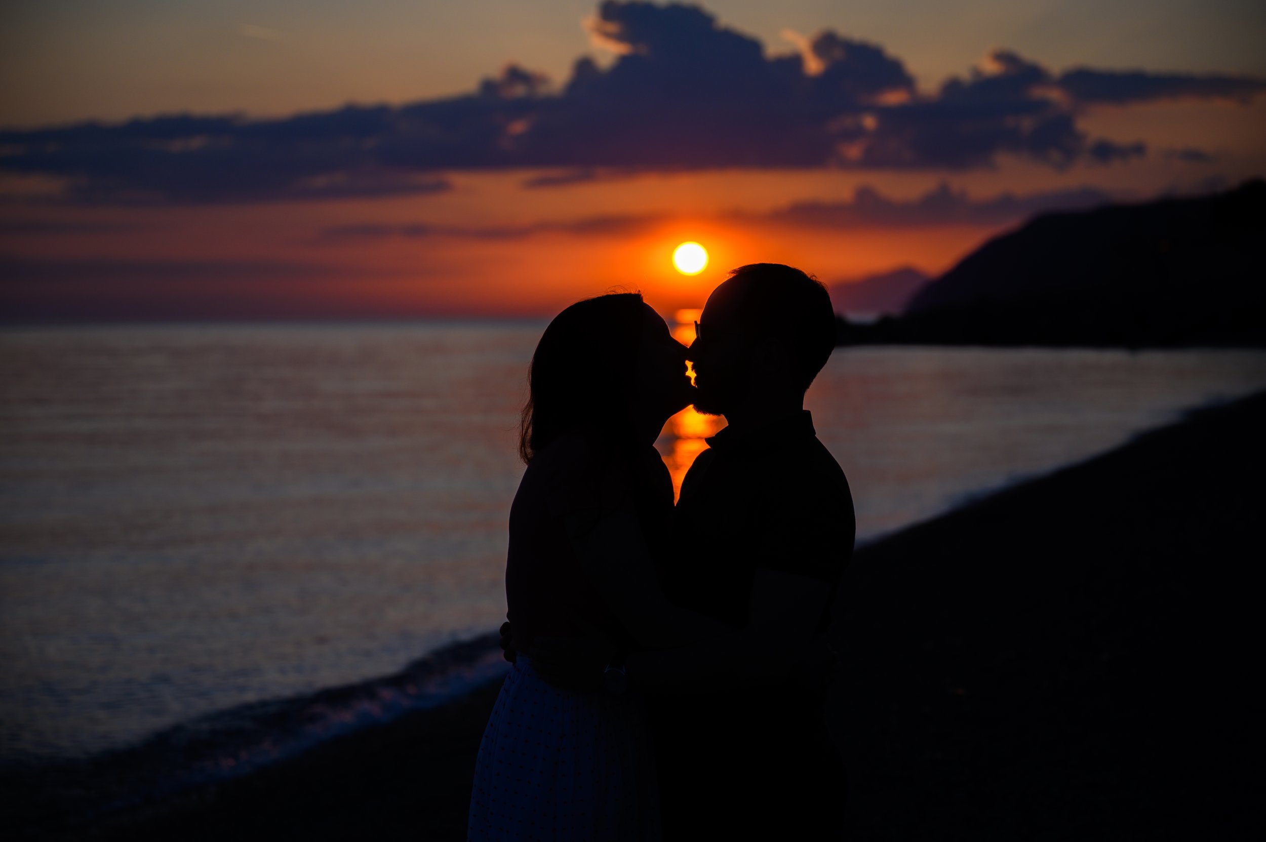 Ein Paar küsst sich bei Sonnenuntergang am Strand, Silhouetten vor einem orange-roten Himmel mit Wolken und Bergen im Hintergrund.