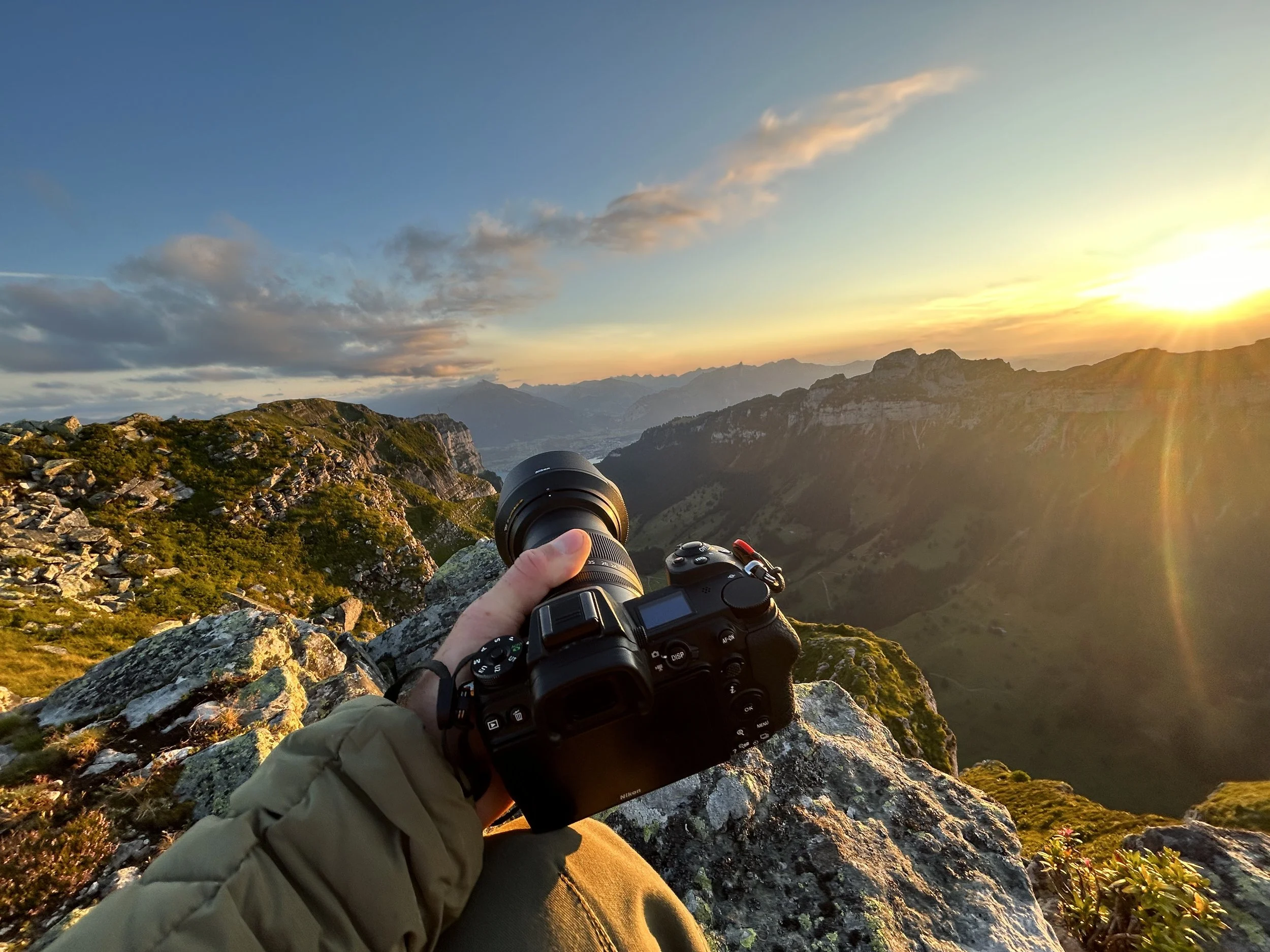 Fotograf, der auf einem Berggipfel sitzt und eine Kamera hält, während er eine malerische Sonnenuntergangsszene über einer grünen Berglandschaft fotografiert.