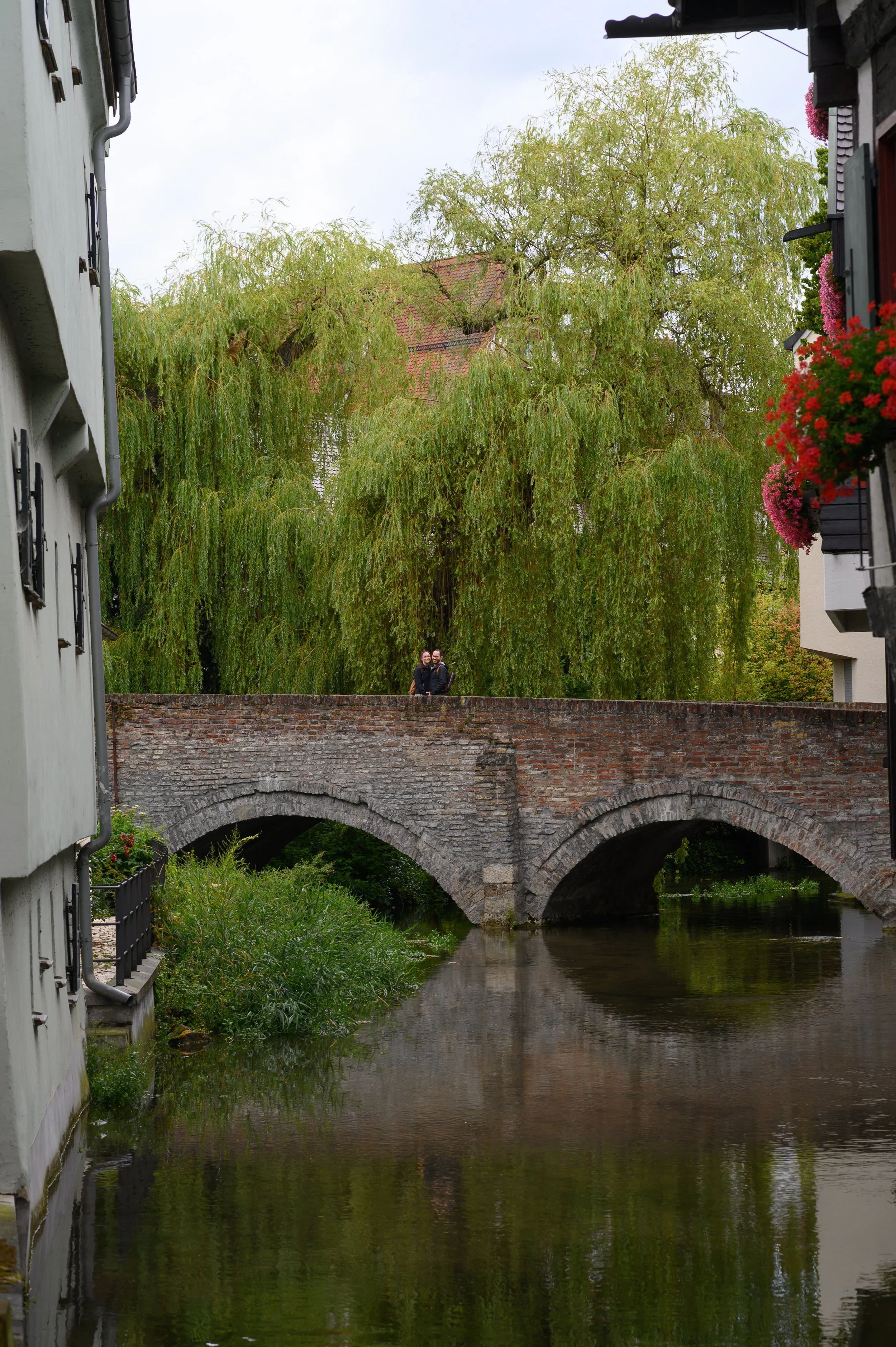 Ein steinerne Brücke über einen Fluss mit zwei Menschen darauf, umgeben von grünen Bäumen und blühenden Pflanzen an den Gebäuden.
