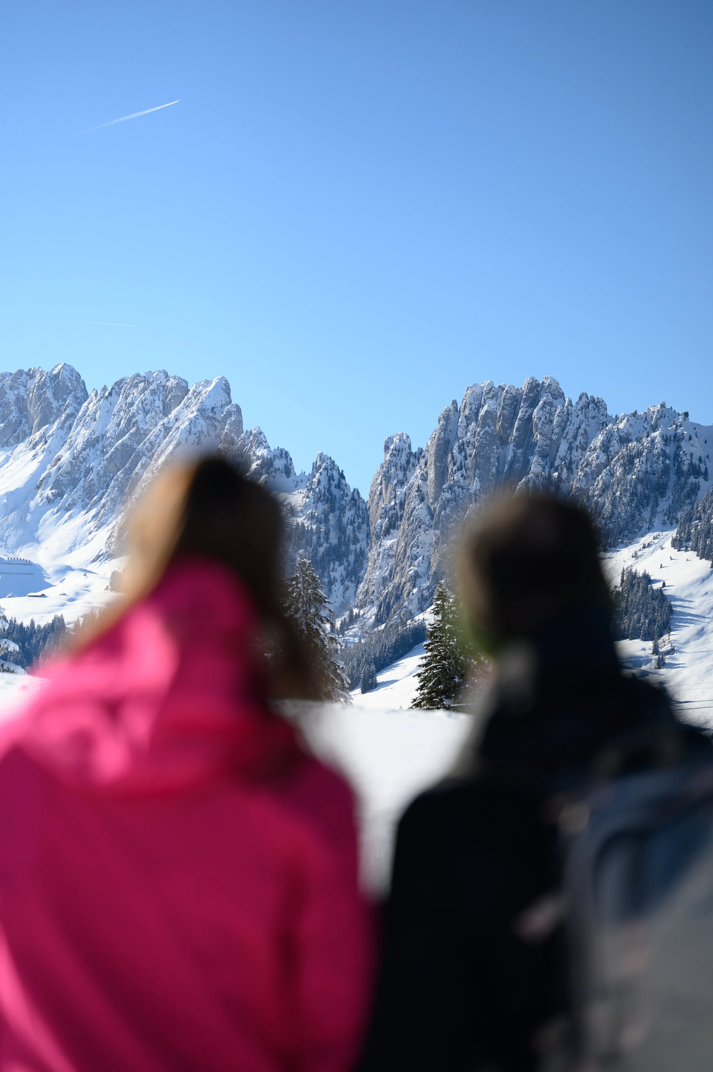 Zwei Personen in Winterkleidung betrachten die schneebedeckten Berge in der Ferne unter klarem blauen Himmel.