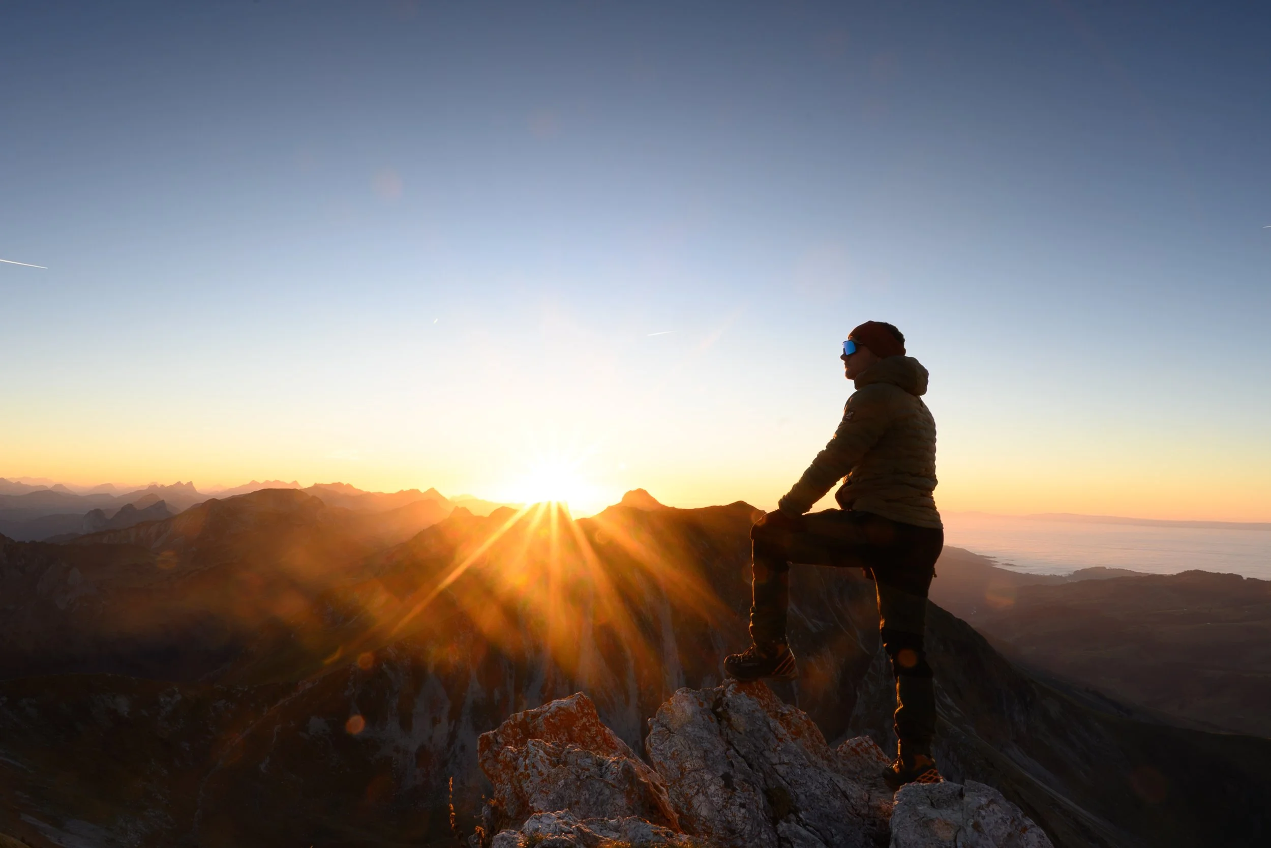 Ein Bergsteiger sitzt auf einem Felsen und beobachtet den Sonnenaufgang über den Bergen bei Sonnenuntergang.