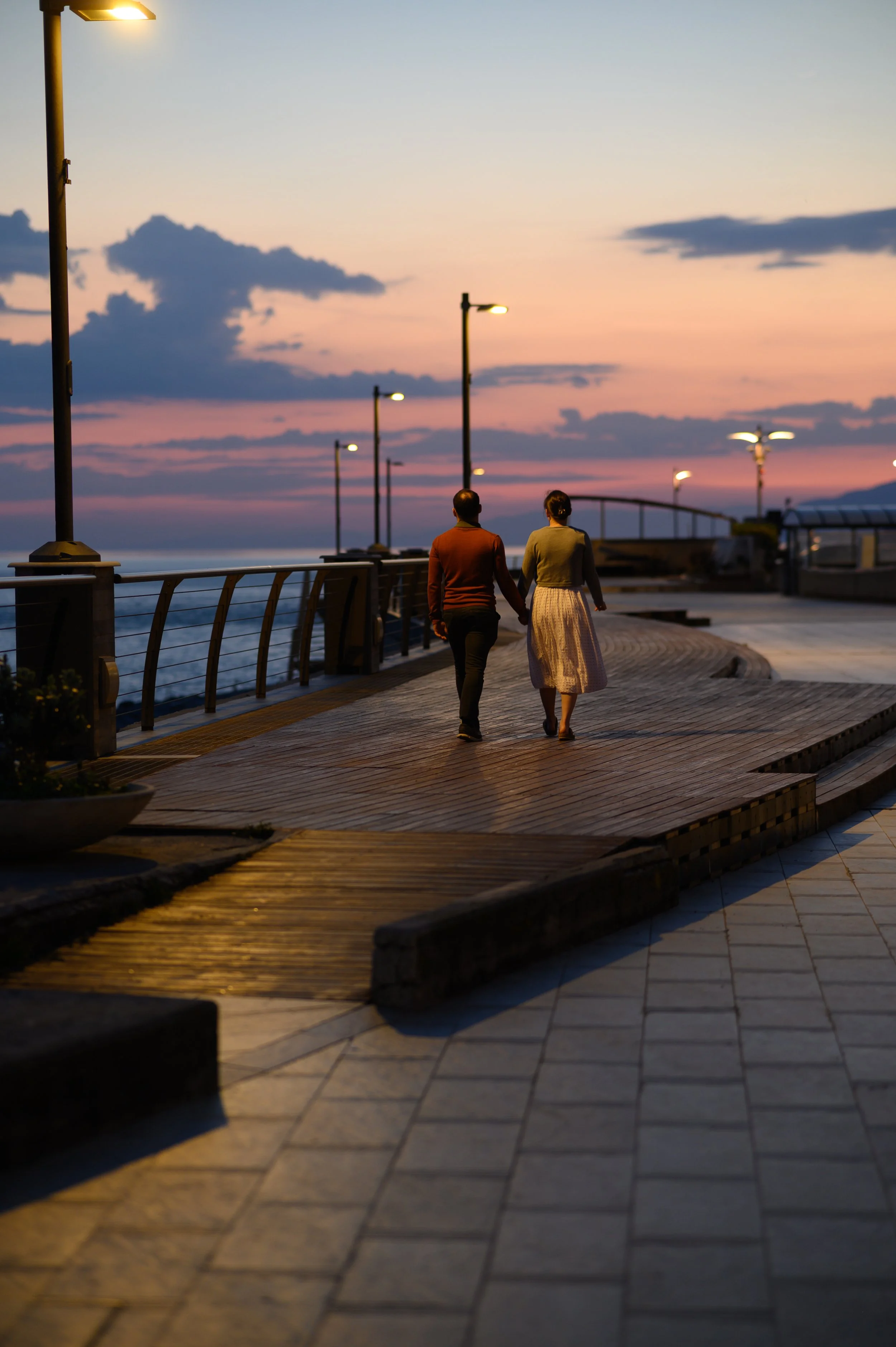 Ein Mann und eine Frau gehen bei Sonnenuntergang Hand in Hand entlang einer Uferpromenade.