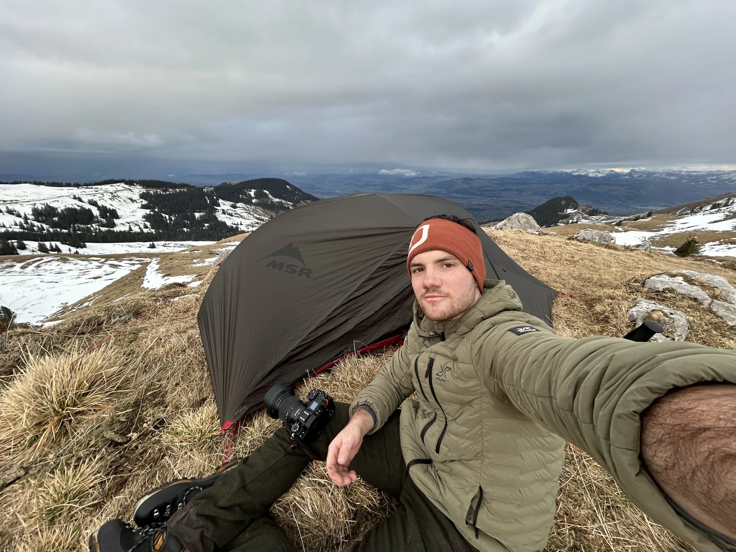 Ein Mann sitzt vor seinem Zelt auf einer Bergwiese, umgeben von schneebedeckten Hügeln, mit einem Berg in der Ferne und dunklen Wolken am Himmel.