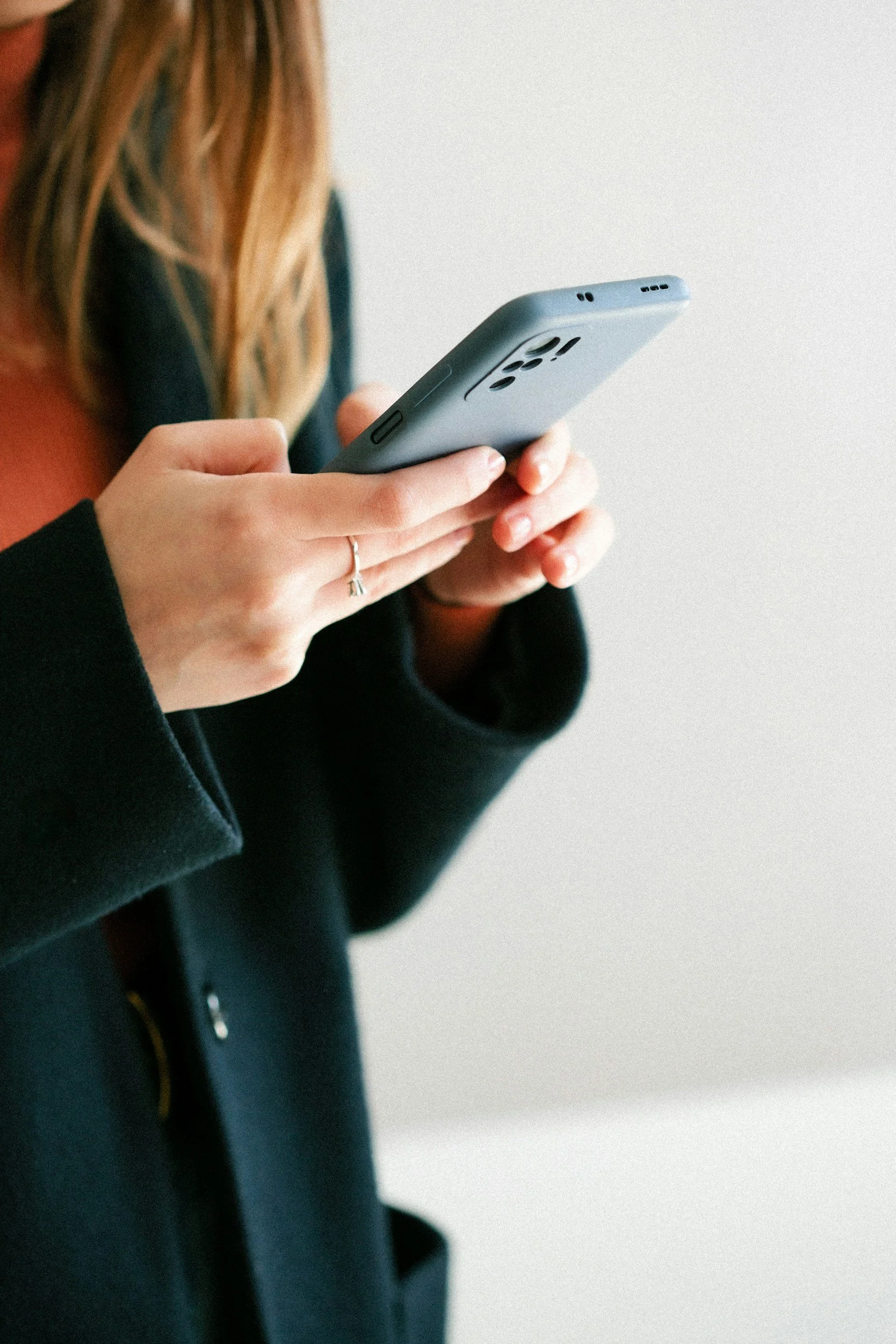 Person holding a smartphone with both hands, wearing a black coat and a ring on their finger, against a plain background.
