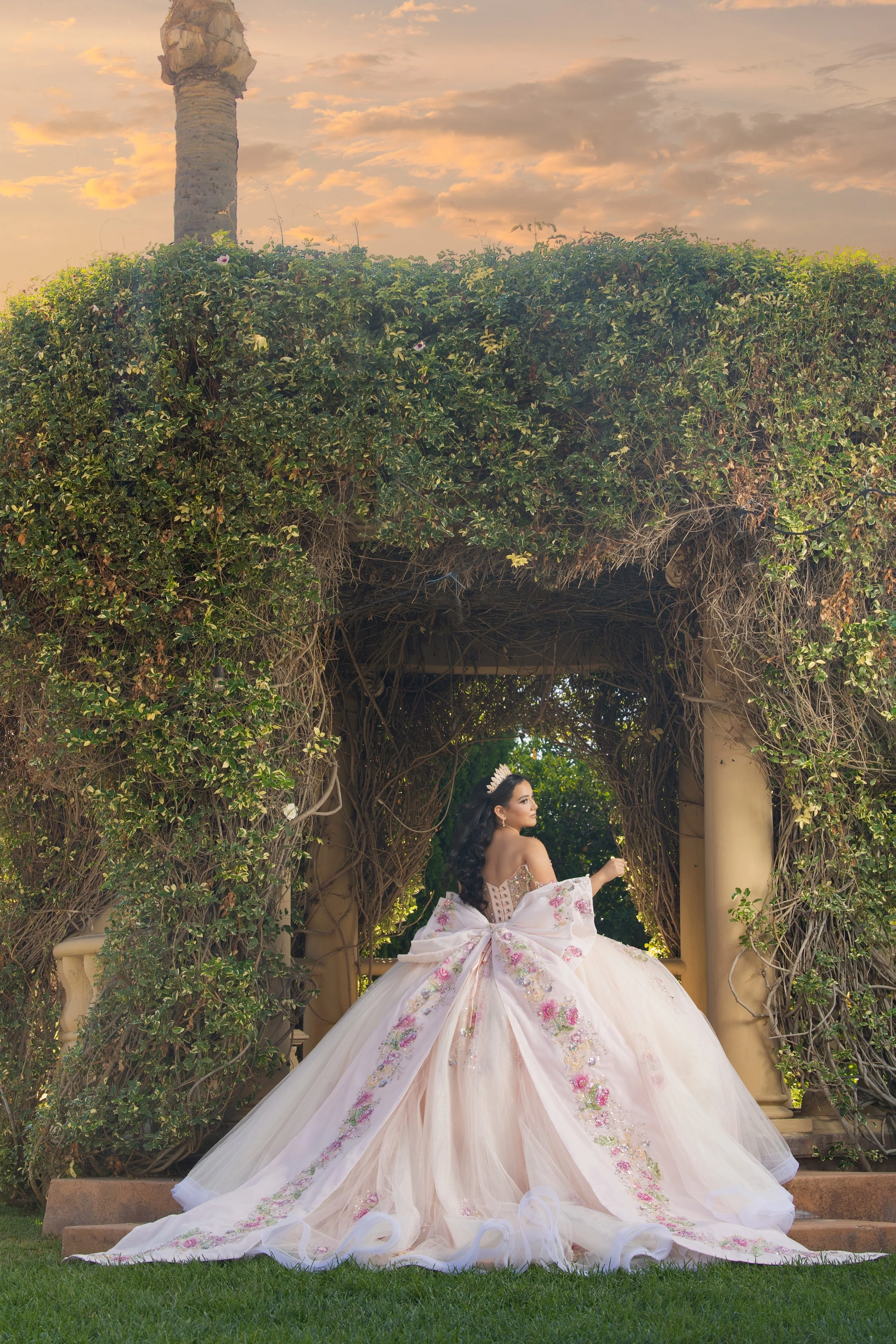 A woman wearing a ball gown and tiara stands in front of a vine-covered archway at sunset for her pre-photoshoot at Rancho Jalisco in Redlands, Riverside, in the IE.