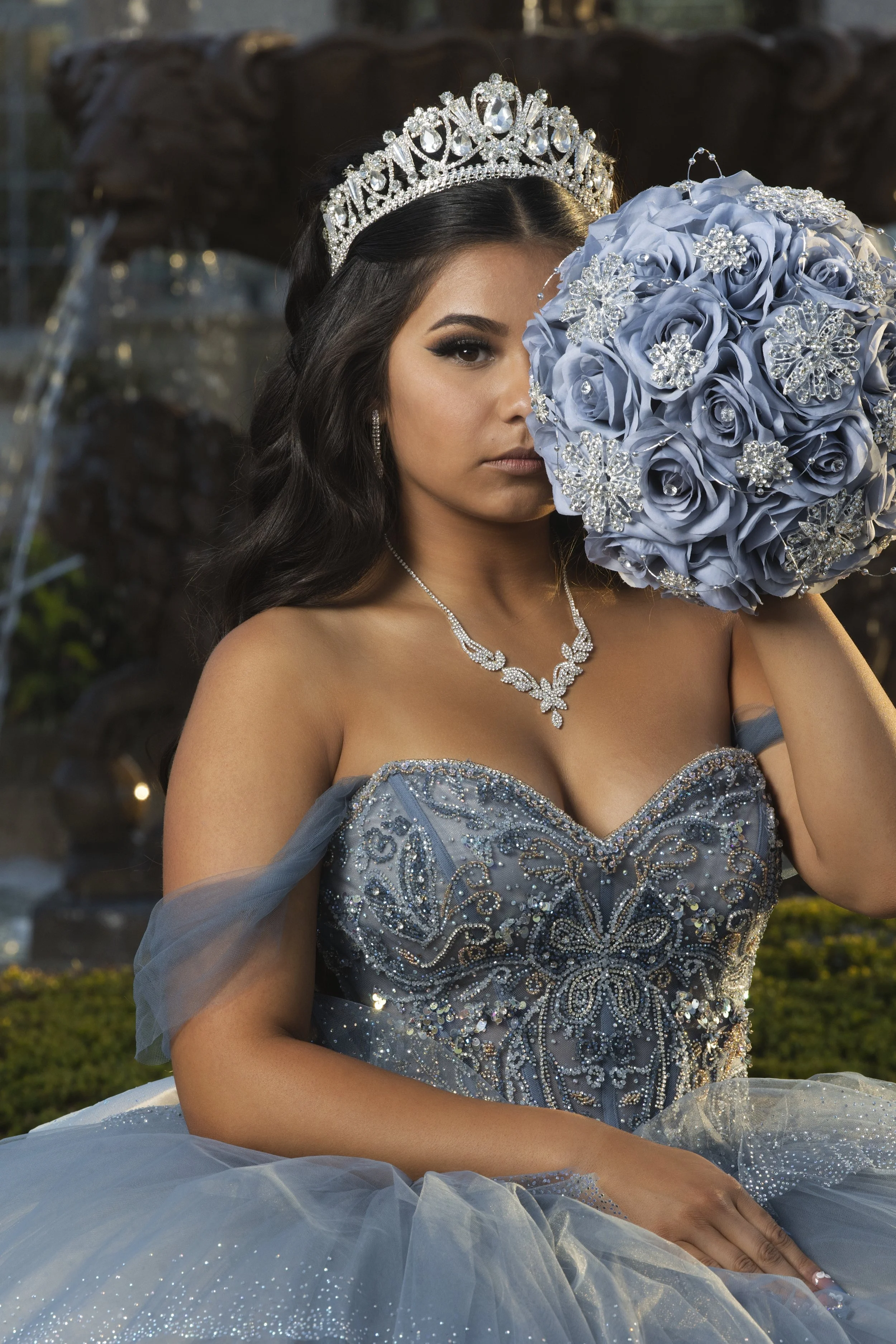 Young woman in an elegant, embroidered blue gown with jewelry, holding a bouquet of blue roses near her face, wearing a silver crown with the word 'Queen' in rhinestones, outdoors near a fountain. Prephotoshoot at Rancho Jalisco, Redlands Inland.