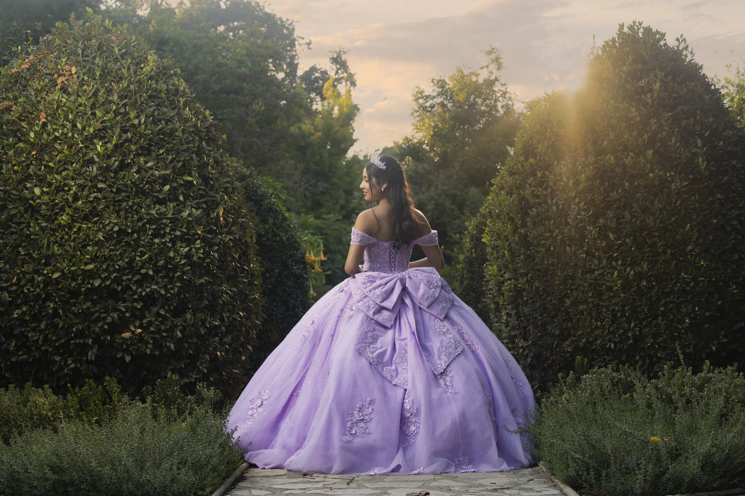 A woman in a lavender princess-style ball gown with lace embroidery and a large bow on the back, standing on a stone pathway between lush bushes, with trees and a sunset sky in the background at Los Angeles Arboretum.