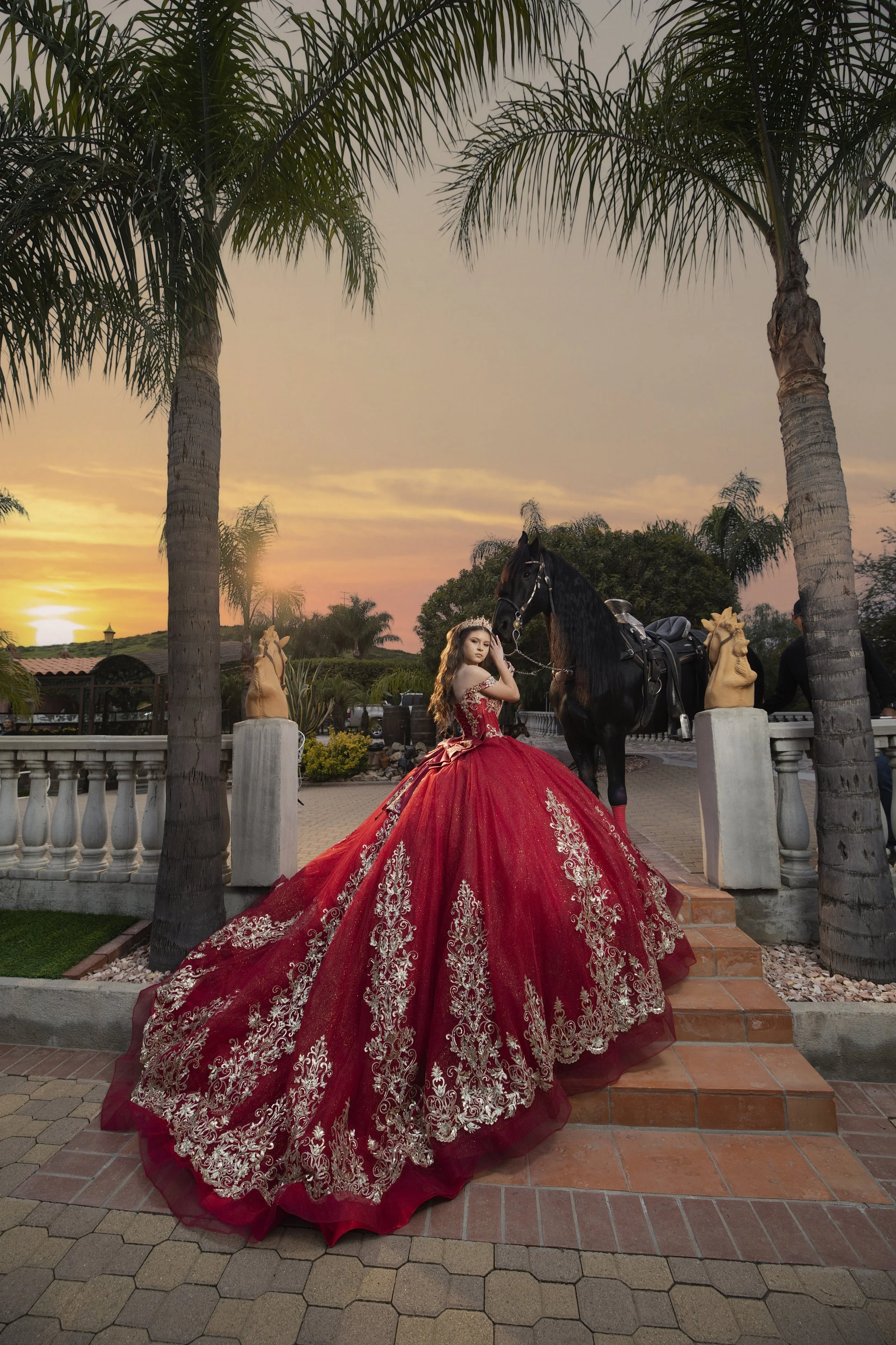 A young woman in an ornate red ball gown with gold embroidery stands on steps next to a black horse at sunset in an outdoor setting with palm trees and stone statues with Rancho El Escondido and their Friesian horse.