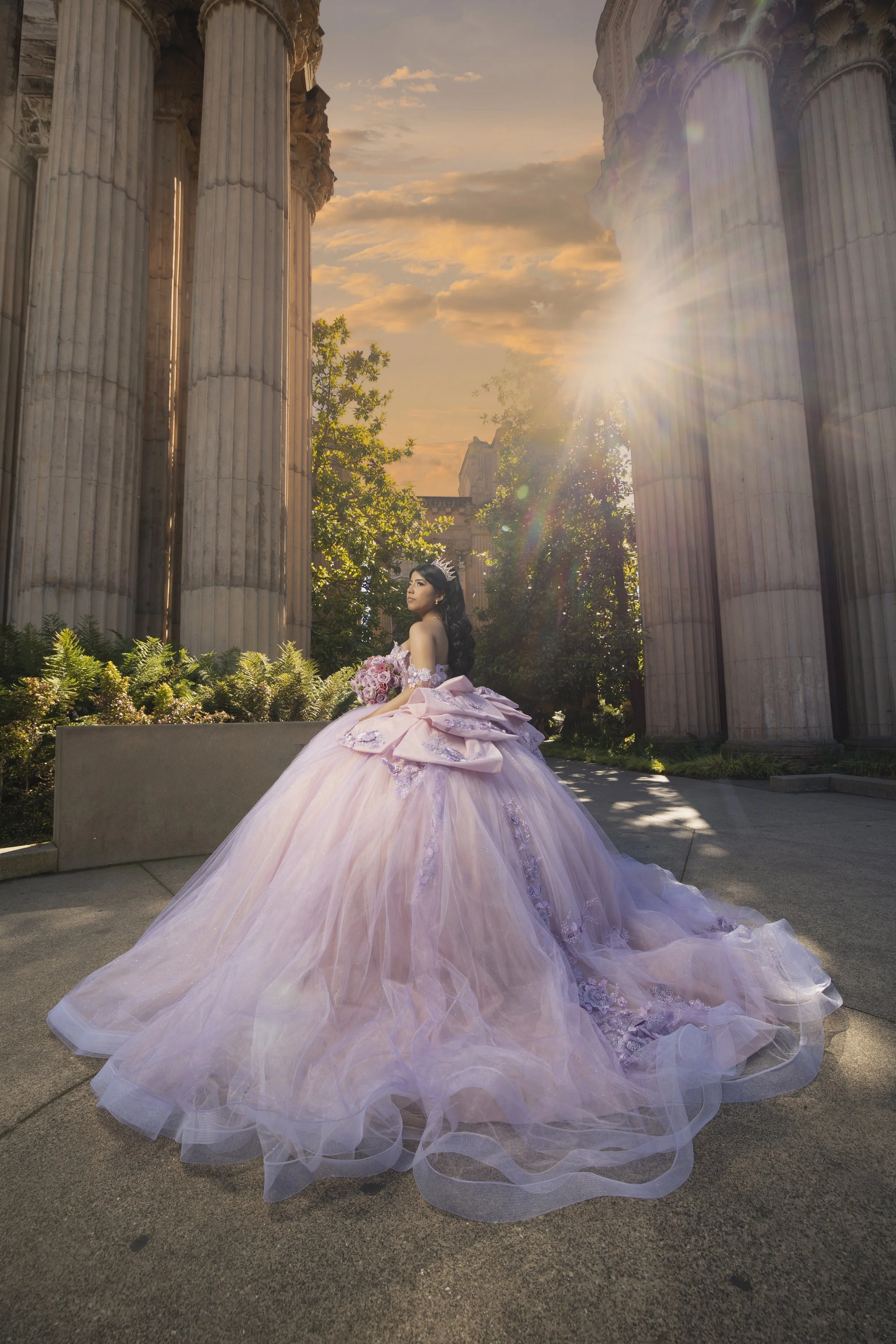 A woman dressed in a pink ball gown with ruffles and lace details, holding a bouquet of pink roses, standing outdoors near ancient columns at sunset in San Francisco at the Palace of Fine Arts known for it's lake and Italian style architecture. 