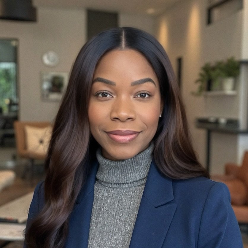 A woman with long dark hair, wearing a gray turtleneck sweater and a navy blazer, smiling in a well-lit modern interior with furniture and plants in the background.