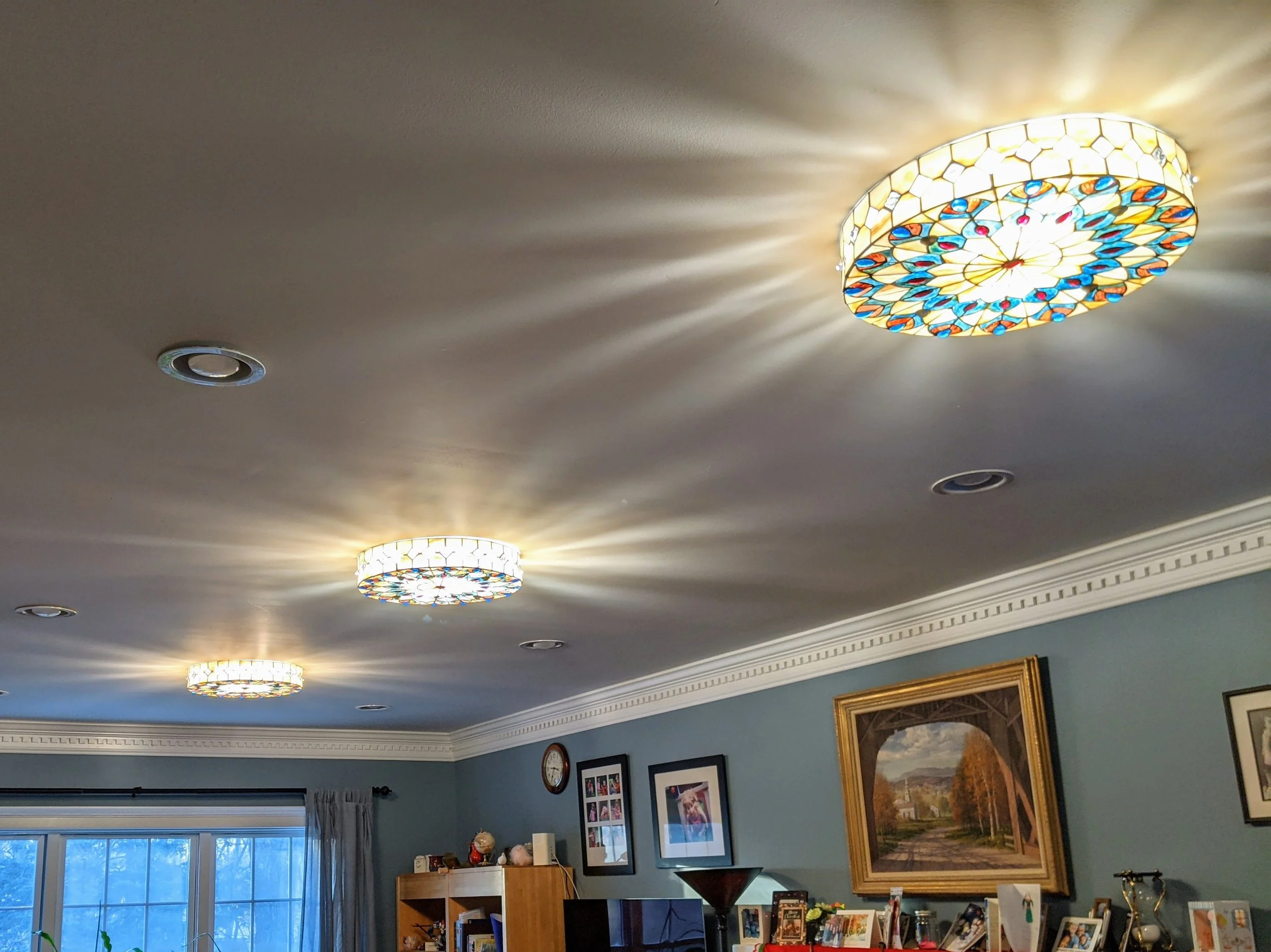 Ceiling with three stained glass LED light fixtures casting colorful light and shadows, in a living room with framed pictures and art on blue walls.