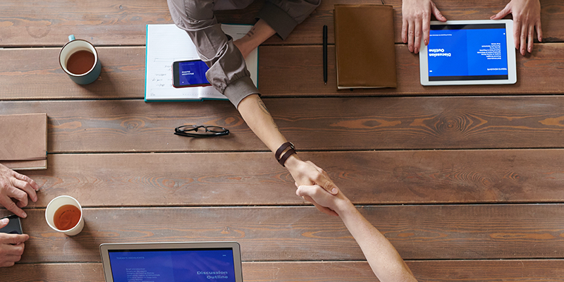 Two people shaking hands across a wooden conference table with laptops, notebooks, glasses, and cups of coffee.